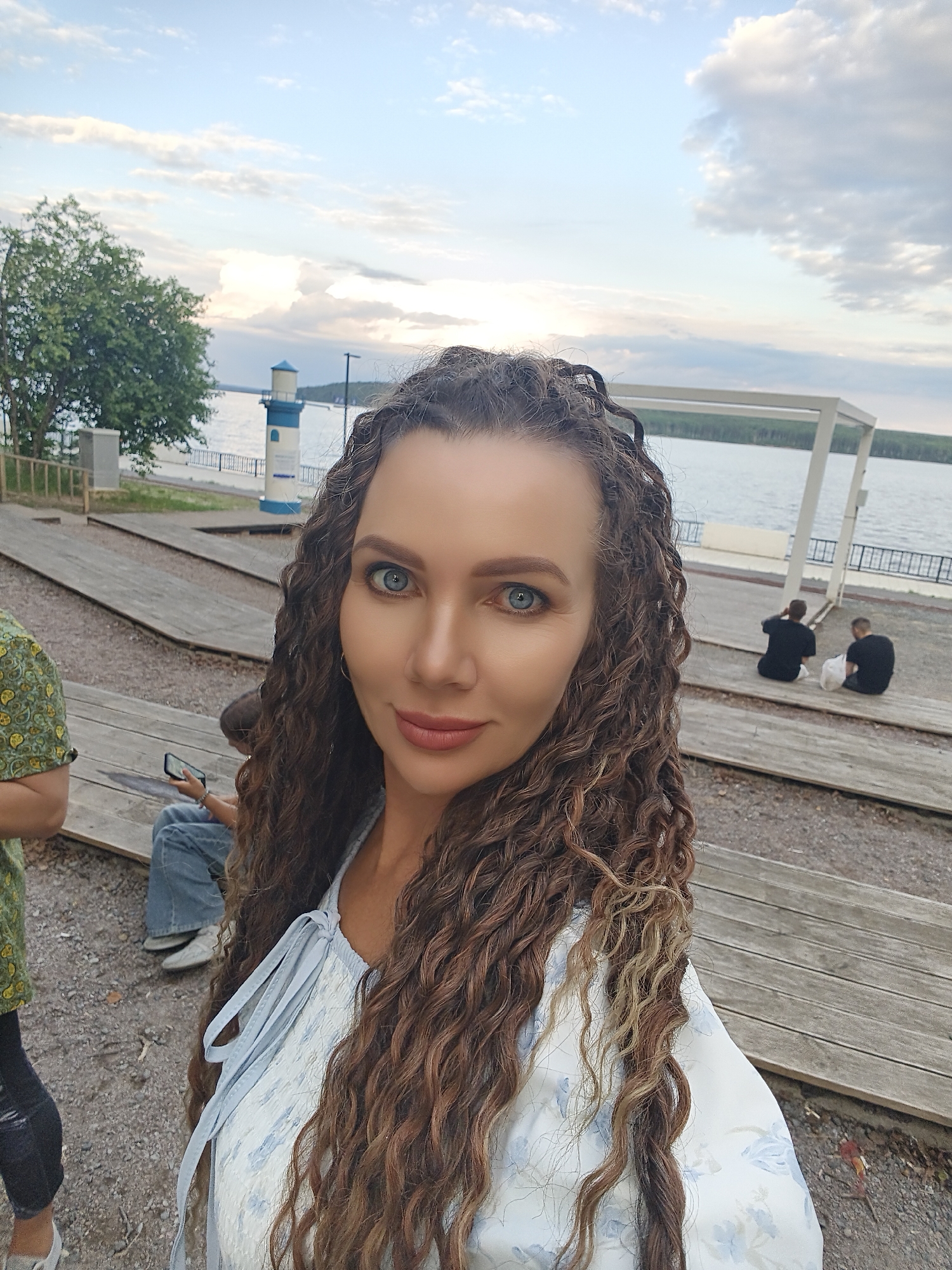 This is a selfie taken by a woman with long, curly brown hair and blue eyes. She is wearing a light blue dress with a floral pattern. She is smiling at the camera.
The background shows a waterfront scene with a lake, a lighthouse, a tree, and wooden bleachers. Two men in black shirts are sitting on the bleachers in the background. The sky is partly cloudy, suggesting it is late afternoon or early evening. The setting appears to be a public park or recreational area.
There is no visible text in the image. The overall mood is relaxed and pleasant, with the woman appearing happy and comfortable. The location context is Chaikovsky, Russia.