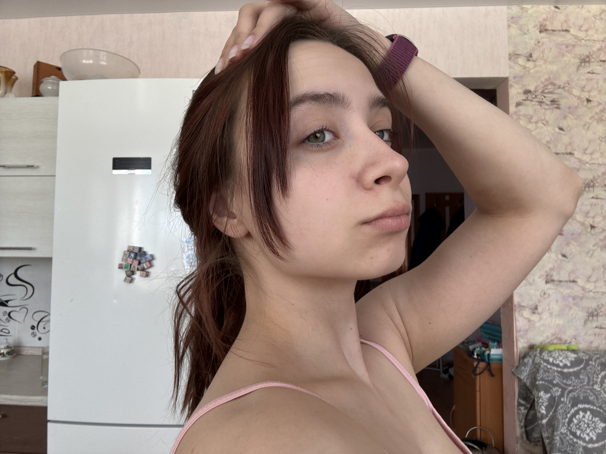 A young woman with auburn hair poses indoors. She is wearing a pink tank top and a fitness tracker on her wrist. Her right arm is raised, with her hand touching her head. She has a slight frown. Behind her, a white refrigerator is visible, adorned with colorful magnets and decorative stickers. In the background, a patterned wallpaper can be seen along with a glimpse of furniture in what appears to be a bedroom. There is no explicit evidence in the image to confirm the location. The model's information indicates that the location may be in Прикамский, Russia.