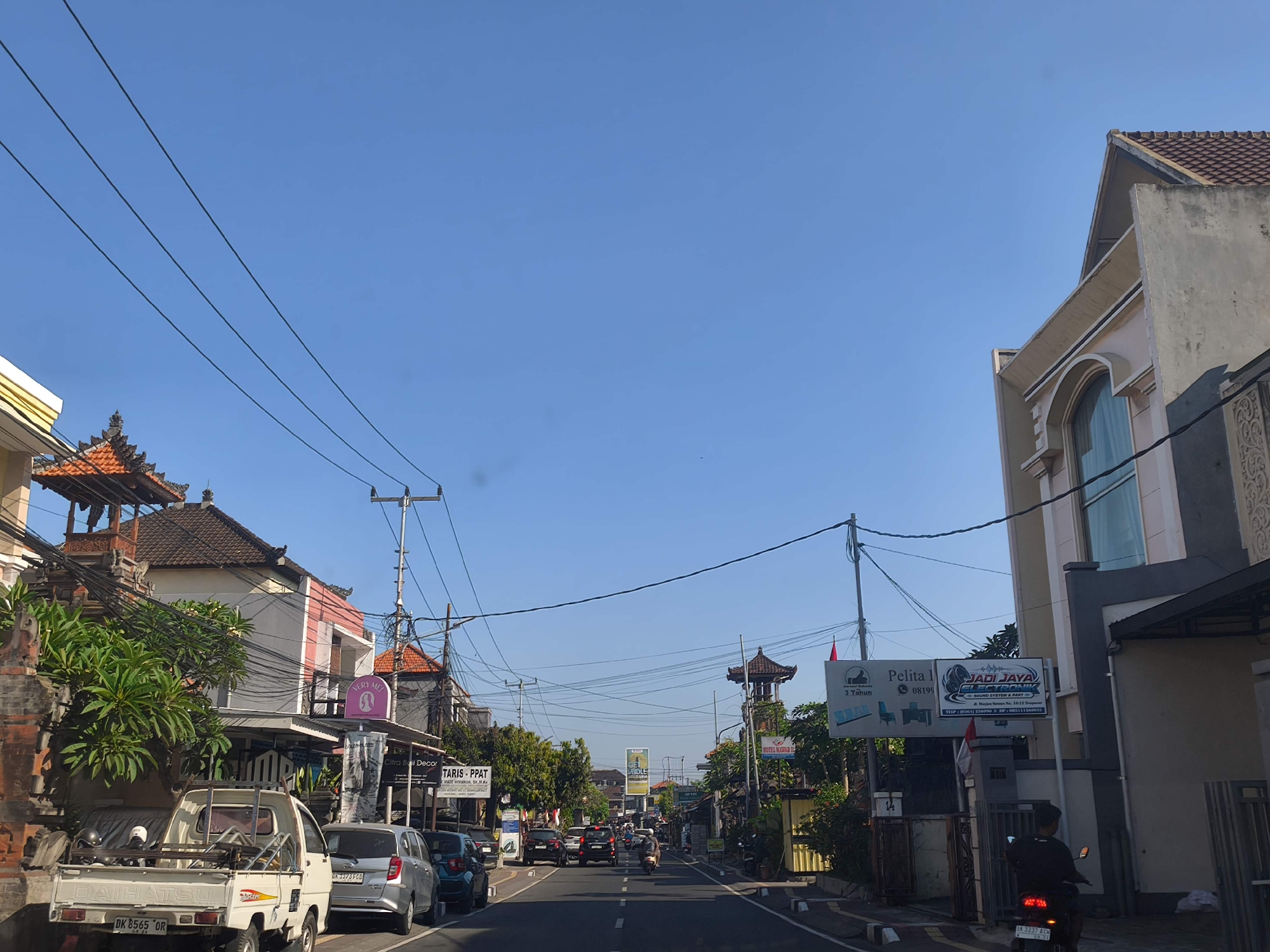 The image depicts a street scene in Denpasar, Indonesia, on a clear day with a blue sky. The perspective is from a vehicle traveling down a road lined with buildings and vegetation. On the left side of the road, there is a white pickup truck with the license plate "DK 8565 QR". Adjacent to the truck is a building with traditional Balinese architectural elements, including a tiled roof and ornate carvings. Further down the street, there are various shops and businesses, some with signs in Indonesian. One sign reads "TARIS - PPAT" and another "Pelita Jaya Sound System & Part". Several cars and a motorcycle are visible on the road, indicating moderate traffic. The right side of the road features a multi-story building with a large arched window and a sign for "JADI JAYA SOUND SYSTEM & PART". Overhead, numerous power lines crisscross the sky. The overall scene is characteristic of a commercial and residential area in a Southeast Asian city.