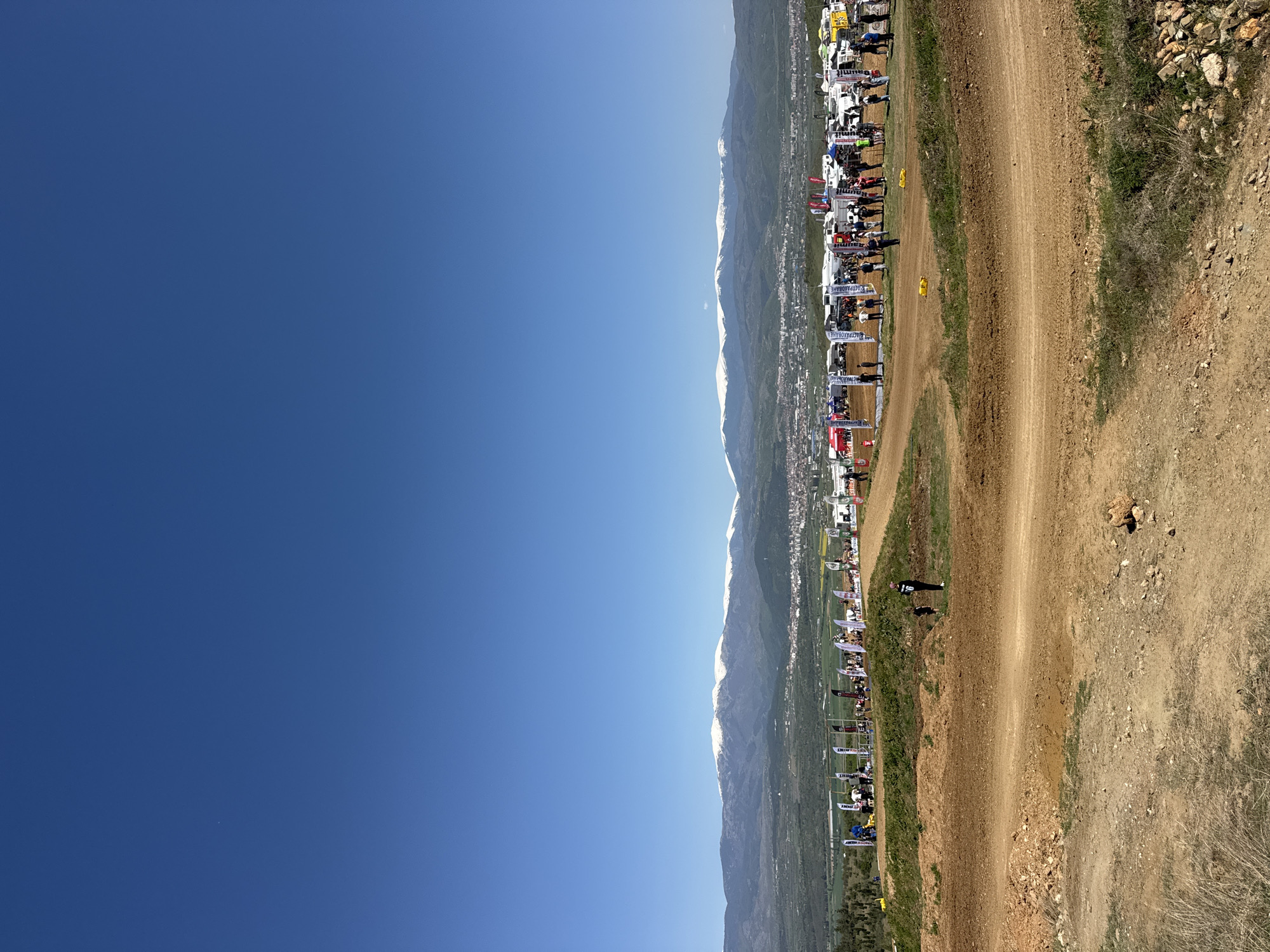 The image depicts a bright, clear day in Selishte, Bulgaria. A dirt track runs vertically through the right side of the image, bordered by a grassy area on the left. The background features snow-capped mountains, a city landscape, and a blue sky that dominates the left side of the frame. Along the left edge of the track are numerous tents, vehicles, and people, suggesting an event or competition is taking place. A single person is visible on the dirt track itself. The ground to the right of the track is rough terrain with vegetation.