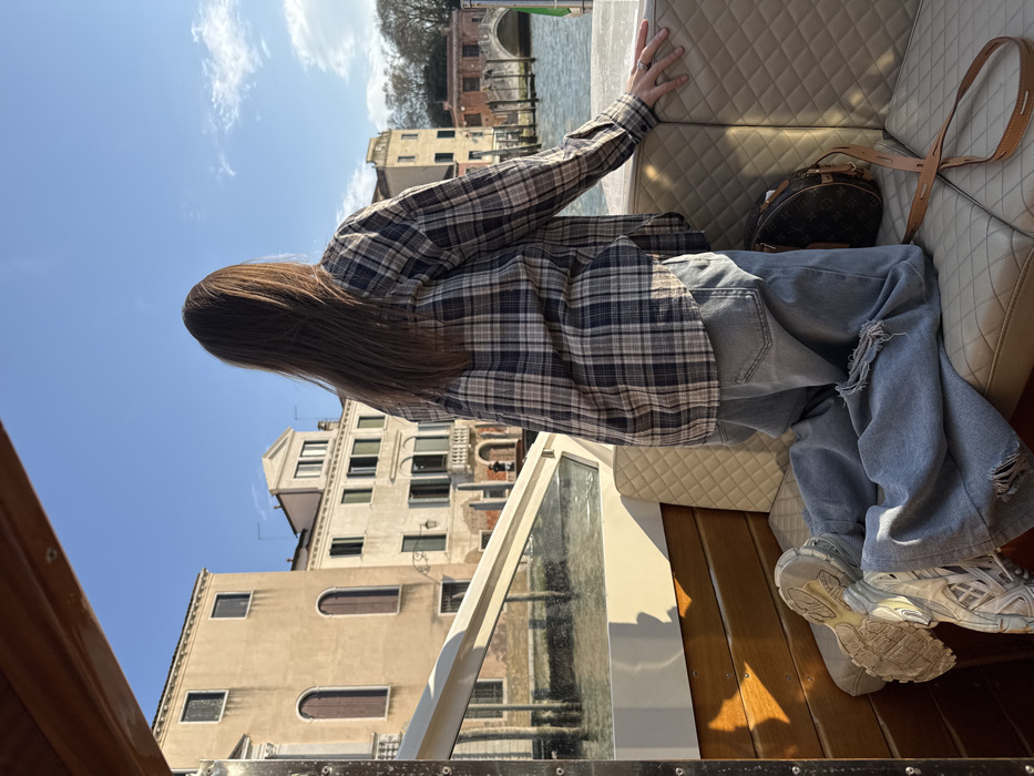 The image depicts a person kneeling inside a boat on a canal in Venice, Italy, viewed from behind under a clear blue sky. The individual has long brown hair and is wearing a blue, black, and beige plaid button-up shirt, light-wash denim jeans with visible rips, and light-colored chunky athletic shoes. They are positioned on a light-cream diamond-quilted upholstered seat, with their left hand resting on the boat's upper rail. A brown monogrammed designer handbag with a tan leather strap is placed on the seat beside them. The boat's interior features wooden paneling and a clear glass windshield. Beyond the boat, historic multi-story buildings, characteristic of Venetian architecture with varied facades, including arched and rectangular windows, line the canal. An arched stone bridge is visible in the distance across the waterway. The scene is illuminated by bright sunlight, casting distinct shadows.