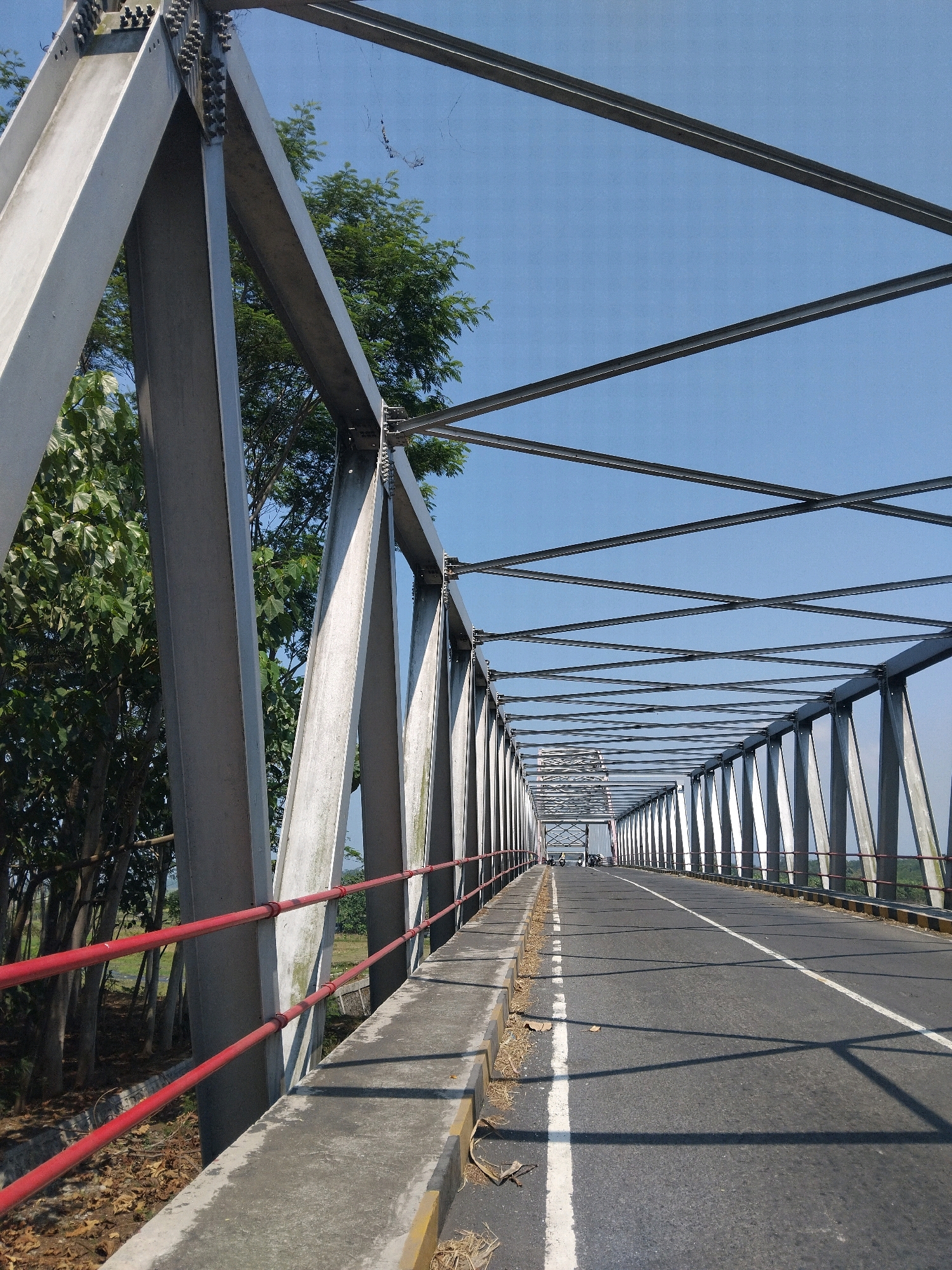 This is a ground-level, medium-angle shot of a steel truss bridge over a road in Selokanayar, Indonesia. The bridge has a gray metal structure with a dark asphalt road running across it. The road has a white dividing line and is bordered by concrete sidewalks and red railings. On the left side of the bridge, there are trees and vegetation. The sky is clear and blue. In the distance, there are a few vehicles on the bridge, including motorcycles.