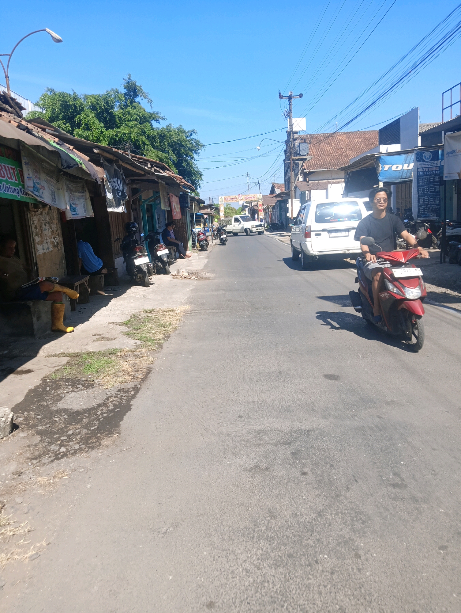 A daytime, eye-level, full shot captures a street scene in Sokaraja, Indonesia. The road, paved with asphalt, is flanked by low-rise buildings and shops on the left. A man in a black t-shirt and patterned shorts is riding a red scooter towards the viewer. He is wearing sunglasses and appears to be looking forward. Behind him, a white SUV is also moving down the road. On the left side of the road, several people are seated outside shops, some appearing to be resting or waiting. Motorcycles are parked along the sidewalk. The sky is clear and blue, with visible power lines crisscrossing the scene. Trees and foliage are visible behind the buildings. The overall impression is of a typical street in a small Indonesian town on a sunny day.