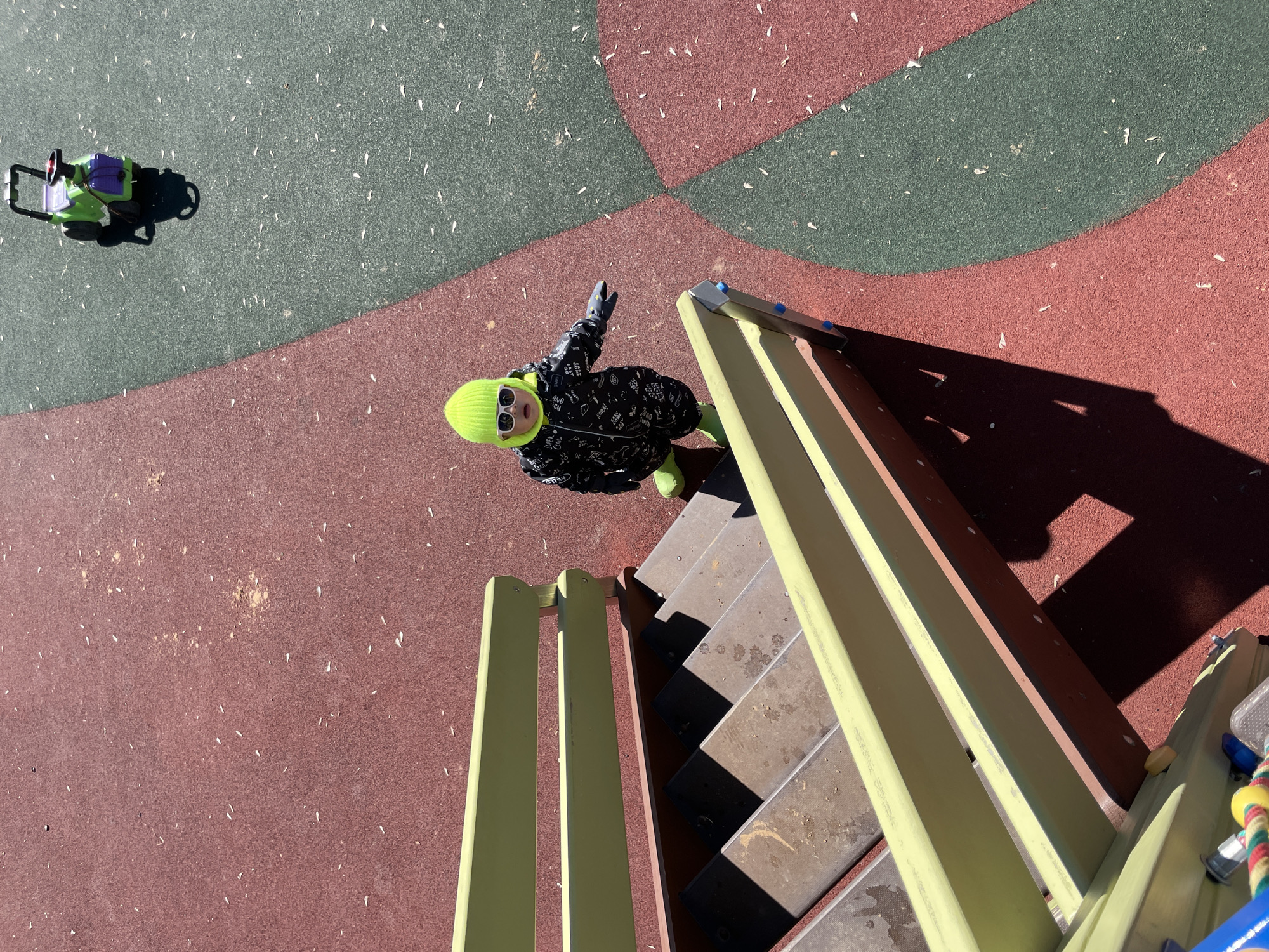 The photograph captures a child on a playground, likely in Izhevsk, Russia. The scene is shot from an elevated perspective, possibly from the top of the play structure. The playground surface is a multi-toned rubber mat in shades of red and green, scattered with small white flecks. A toddler stands near the top of a short set of metal stairs with yellow railings, wearing a dark-colored winter coat patterned with light-colored designs, a neon green hat, neon green boots, and sunglasses. The child's left arm is raised in a wave or gesture. The stairs are weathered, showing signs of wear and shadows cast across the steps. To the left of the child, on the green section of the playground surface, is a green and purple toy. The overall light in the image suggests a sunny day, casting shadows that define the edges of the structures and objects in the scene.