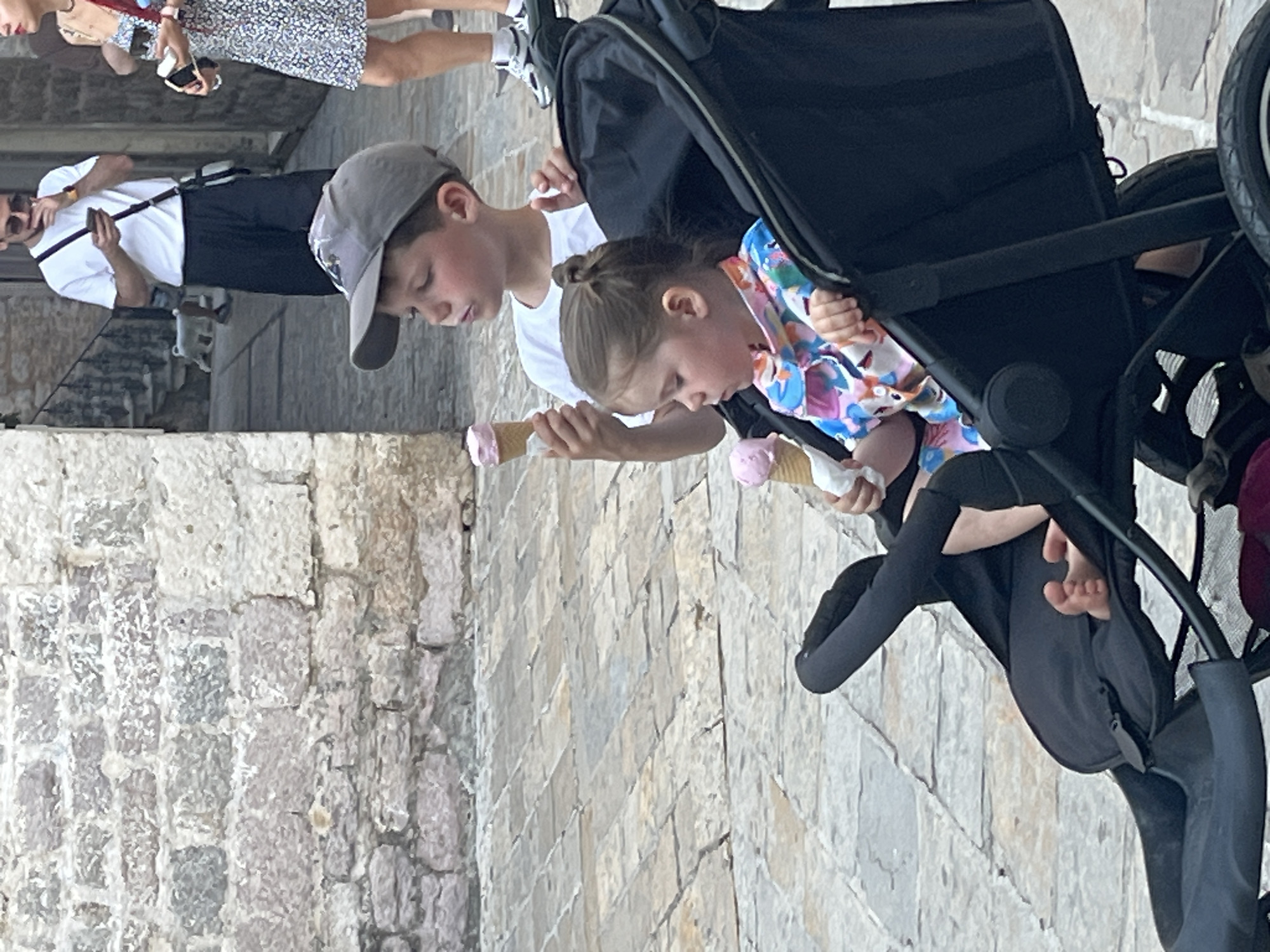 A young boy wearing a grey baseball cap and a white t-shirt holds a pink ice cream cone while standing next to a black stroller. Inside the stroller, a young girl wearing a colorful floral print top sits with her eyes closed, also holding a pink ice cream cone. The scene takes place on a cobblestone street in Budva, Montenegro, with a stone wall visible in the background. Other people are out of focus in the background, and a white dog is walking in the distance.