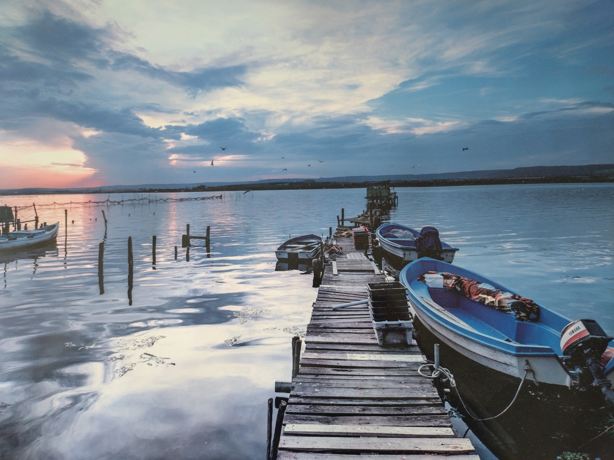 The image depicts a sunset over a body of water, likely a lake or bay, with a weathered wooden dock extending into the water. Several small boats are docked alongside the pier.
The sky is filled with dramatic clouds, the sun setting on the horizon. A line of distant land is visible. Birds are flying above the water. The water reflects the light and clouds, and the dock is made of aged wood. Fishing nets stretched with wooden poles are visible in the left background. A small dilapidated structure stands on stilts at the end of a smaller dock extending out from the main dock.
There are no people visible in the image.
There is no indication that the scene is located in the city of Stara Zagora, Bulgaria.