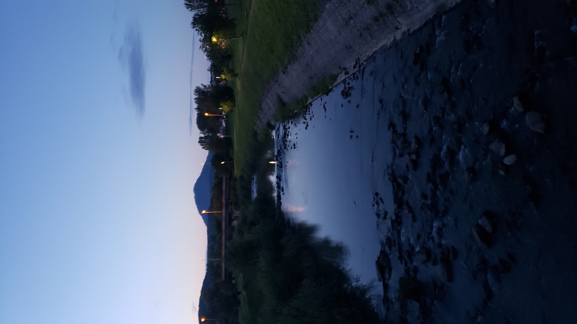 The image shows a view of the Torysa River in Prešov, Slovakia, during twilight. The river flows towards the foreground, with numerous rocks visible in its shallow waters, creating a slightly turbulent surface. The riverbanks are lined with green grass on the right and dense foliage on the left.
In the background, a bridge spans the river. Further beyond, hills are visible, the most prominent being a conical mountain with a peak illuminated by a streetlamp. Several other streetlamps are scattered throughout the scene, casting a warm glow on the grassy areas and the trees. The sky is a deep blue, transitioning to lighter shades near the horizon, with a faint cloud formation. The overall impression is of a peaceful, natural setting within or on the outskirts of the city, likely a park or recreational area. There are no people visible in the image.
