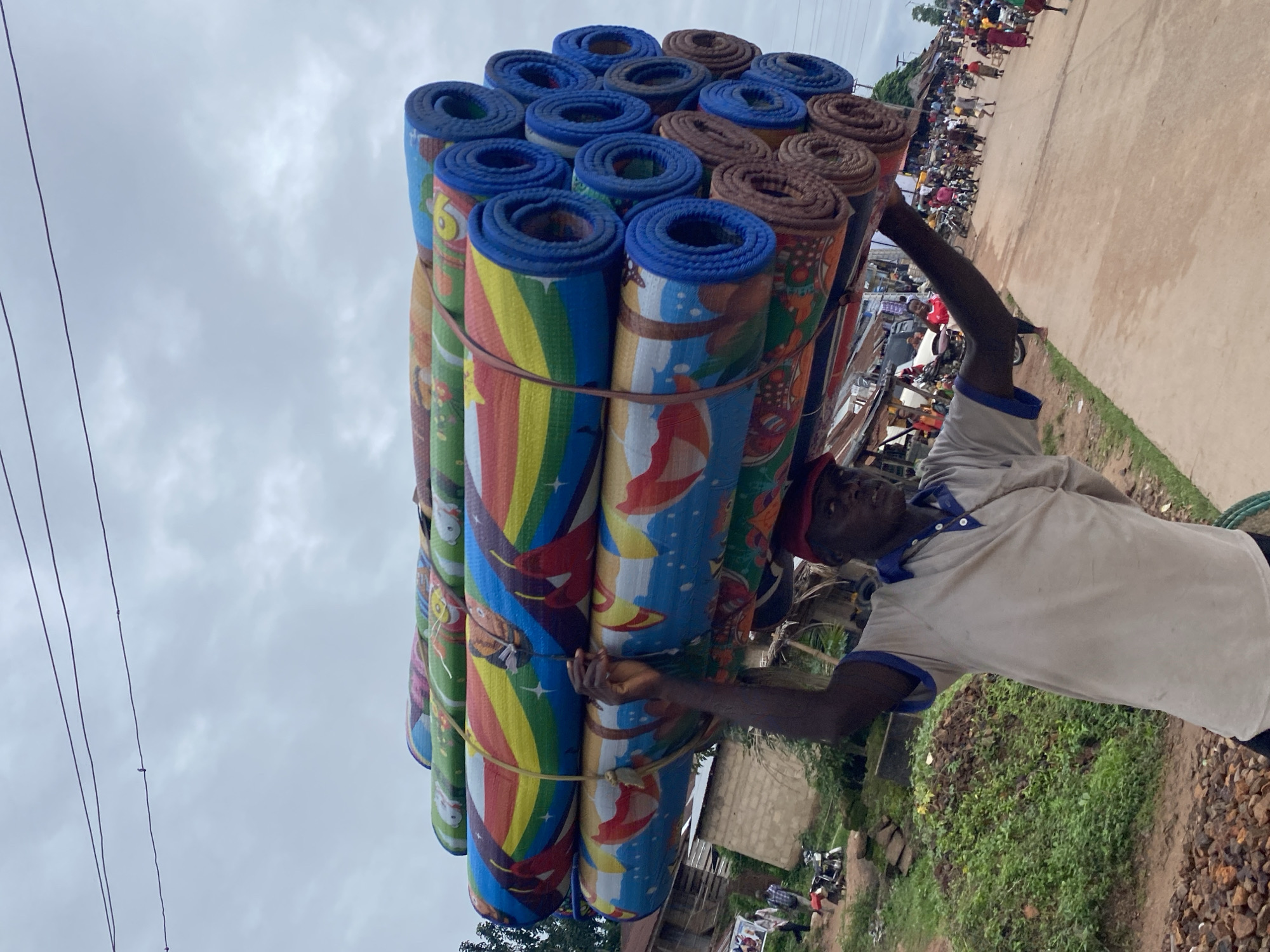 An eye-level shot depicts a man carrying a large bundle of rolled mats on his head and shoulder in what appears to be Igbara, Nigeria. The mats are brightly colored with blues, browns, reds, and yellows. The man is of dark complexion and wearing a light-colored shirt with blue trim. He has his right arm raised as though steadying the mats. In the background is a street with blurred figures of people and motorcycles indicating activity. The surrounding area has vegetation and basic building structures. The sky is overcast.