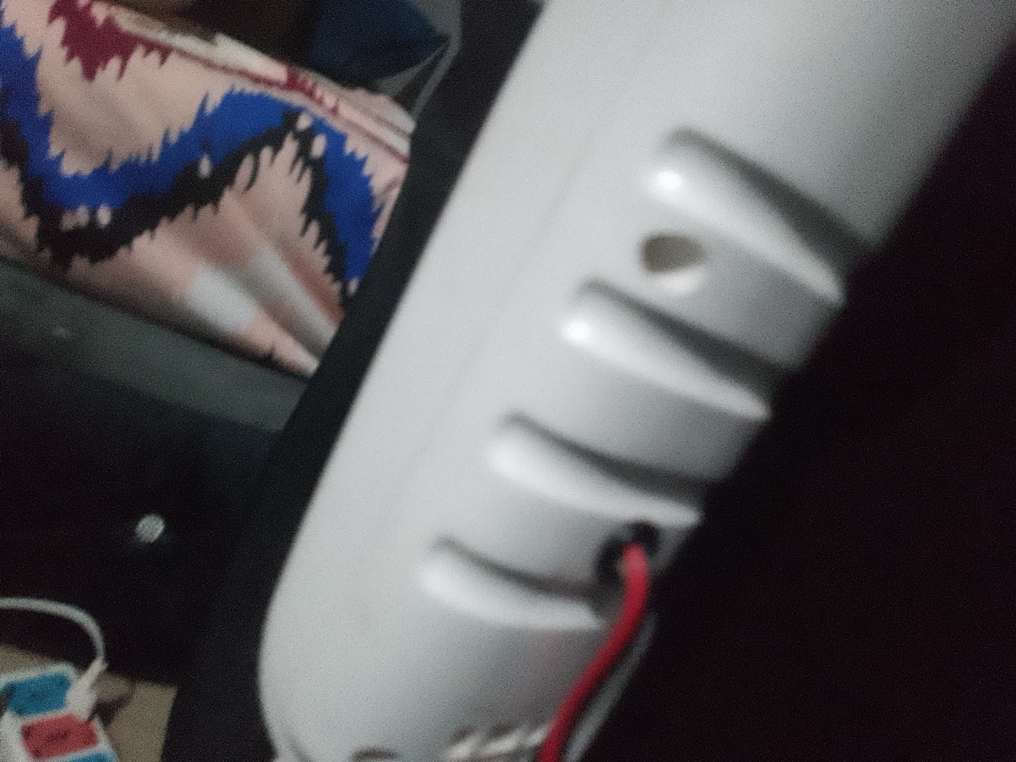 The image shows a close-up, blurry view of a white object with ribbed detailing and a small red wire exiting an opening. In the background, a colorful fabric with a zig-zag pattern is visible, appearing to be a blanket or upholstery. The setting is indoors, with dark shadows suggesting low lighting. No city or specific location details are discernible from the image.