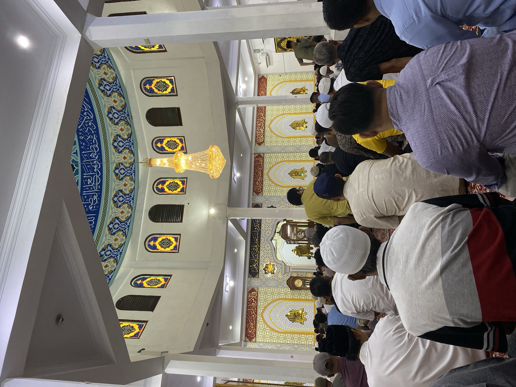 This media file captures a group of men engaged in prayer, likely at a mosque, given the architectural elements. The scene is indoors, with high ceilings, ornate chandeliers, and decorative arched windows featuring stained glass. The walls are adorned with intricate patterns and what appears to be Arabic calligraphy, suggesting a religious or cultural setting in Indonesia, specifically Lhokseumawe.
The subjects are predominantly men, dressed in traditional Islamic attire such as peci (caps) and kurtas or shirts. They are in various stages of prayer, with many in a bowing position (ruku). The atmosphere appears solemn and focused, reflecting the spiritual nature of the activity. The lighting suggests it could be daytime, but the indoor setting makes it difficult to ascertain the exact time or weather conditions outside. The overall impression is one of communal worship and devotion.
The visible text is primarily decorative Arabic calligraphy on the walls, which is common in Islamic architecture and religious spaces. Specific phrases or verses are not discernible due to the resolution and angle.