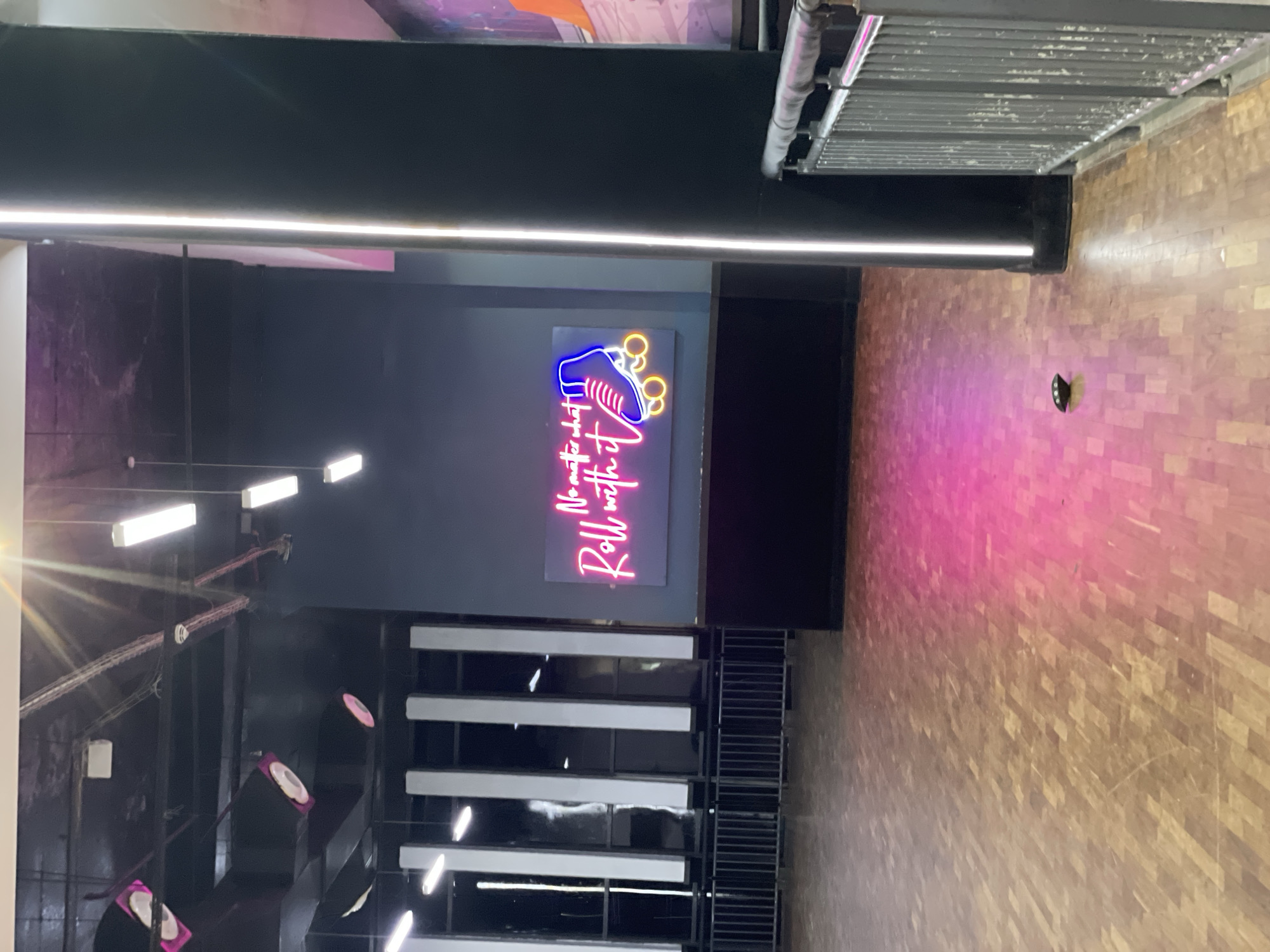 The image depicts the interior of what appears to be a roller skating rink or entertainment venue. The floor is made of polished wood, with areas reflecting pink and yellow light. In the center of the image, a neon sign on a dark blue wall reads "No matter what, Roll with it" and features a graphic of a roller skate. To the right of the neon sign, a tall, black cylindrical pillar with a vertical white light strip is visible. The background shows a dark ceiling with industrial lighting fixtures and what look like decorative pink discs suspended from the ceiling. Metal railings and barriers line the perimeter of the space. A small, dark object with three illuminated points sits on the wooden floor in the lower right quadrant. The scene suggests an indoor recreational facility, possibly in the city of Moba, Nigeria, given the provided context. There are no people visible in the image.