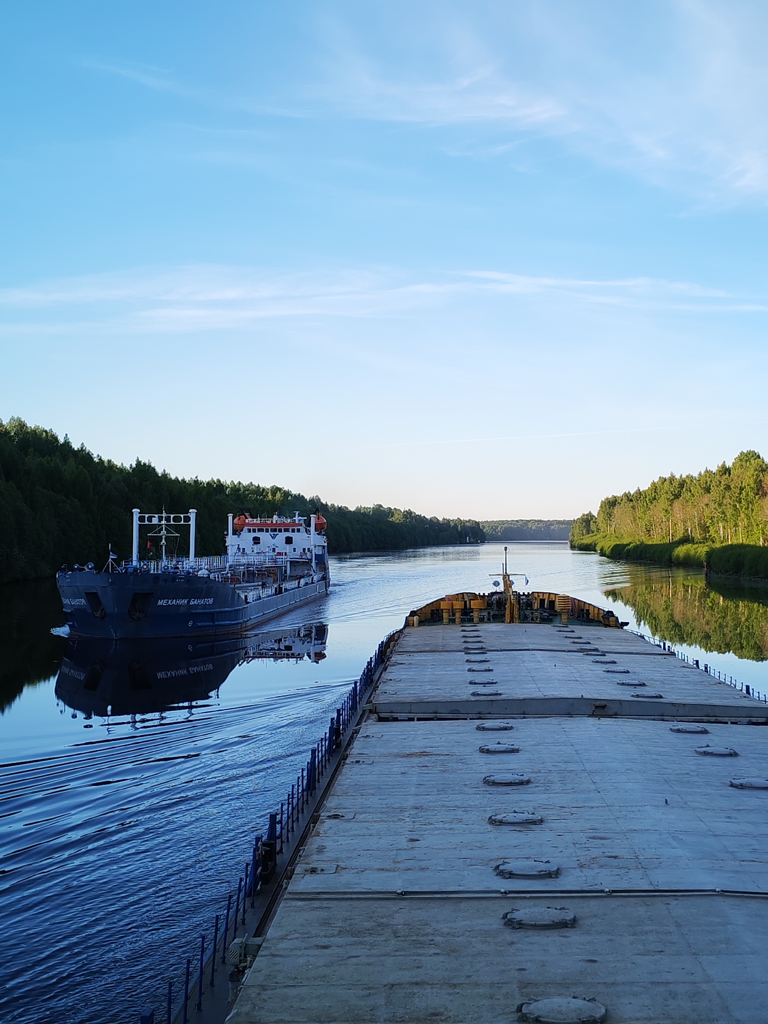 The image captures a peaceful maritime scene on a clear day, likely in the early morning or late afternoon, along a wide river or canal flanked by dense, green forests. The sky above is a bright, clear blue with a few wispy clouds.
The photograph is taken from the deck of a flat-topped barge or cargo vessel, which extends into the foreground and mid-ground on the right side. Its textured deck is light-colored and features multiple circular hatches. Blue railings are visible along the edge of the vessel.
In the mid-ground on the left, a dark blue and white tanker ship is seen moving away from the viewer, passing the vessel from which the photo is taken. The tanker generates a noticeable wake in the calm water. The name "МЕХАНИК БАНАТОВ" (MEKHANIK BANATOV) is clearly visible in white Cyrillic letters on the tanker's hull, with its reflection also visible in the water. The tanker has a multi-deck superstructure with various equipment, including lifeboats or rafts, and antennas.
The water surface is calm, providing clear reflections of both the passing ship and the lush green trees lining the banks. The sun casts a gentle, warm light on the foliage, suggesting a serene atmosphere. No people are visible in the frame. The location context is Старое Петровское, Russia, implying this scene takes place in a waterway within that region.