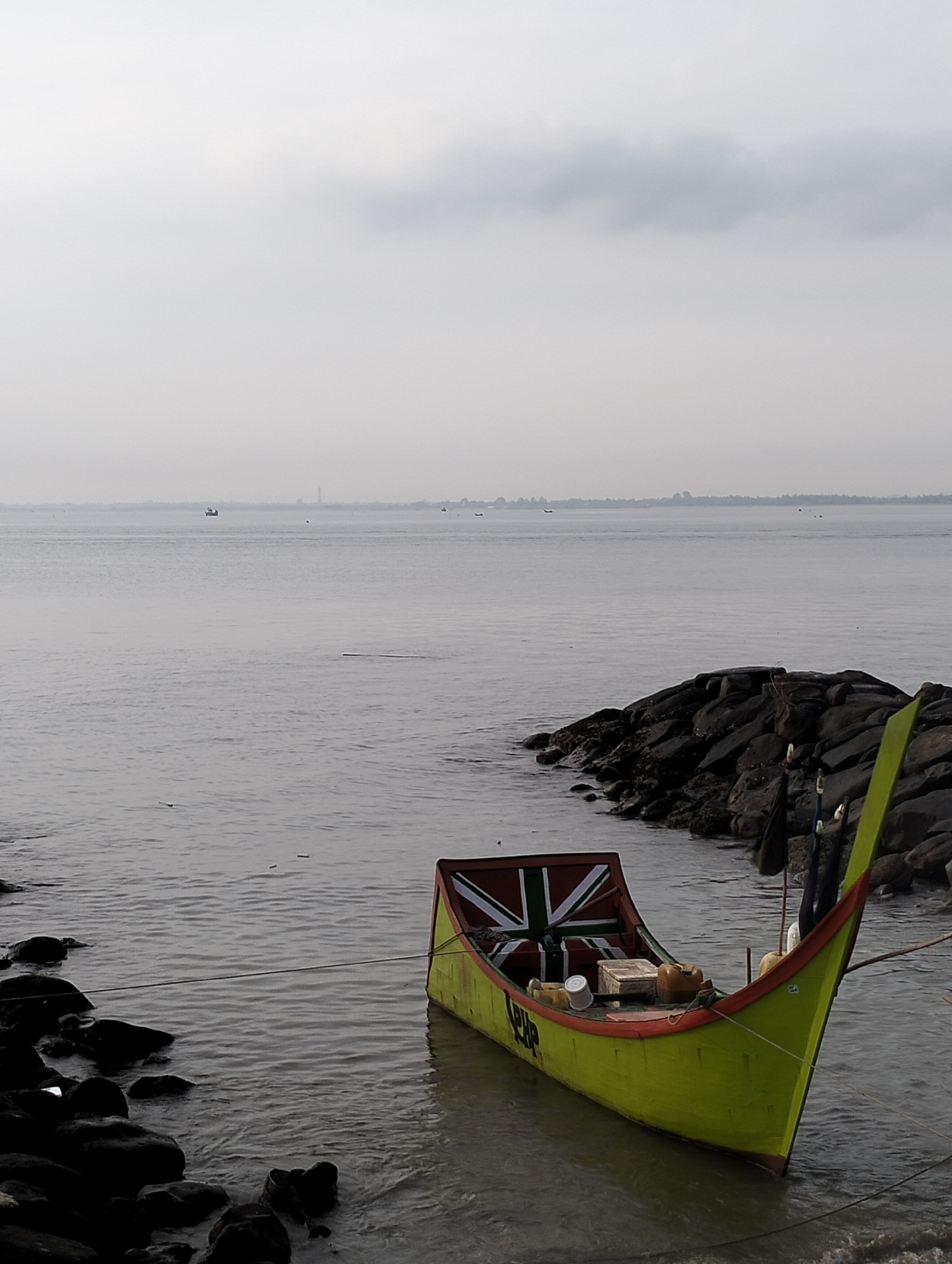A yellow and red trim boat is docked along a rocky shore, partially in the water on a hazy day. The boat's stern displays a stylized Union Jack flag. The interior contains various containers and objects, covered with a tarp. To the right of the boat, a rocky outcrop extends into the water, suggesting a breakwater or shoreline protection. Across the water, a distant, hazy shoreline is visible, punctuated by a few indistinct structures. Small boats are scattered across the calm water. The sky is overcast. This scene is situated in the city of Lhokseumawe, Indonesia, characterized by its coastal setting.