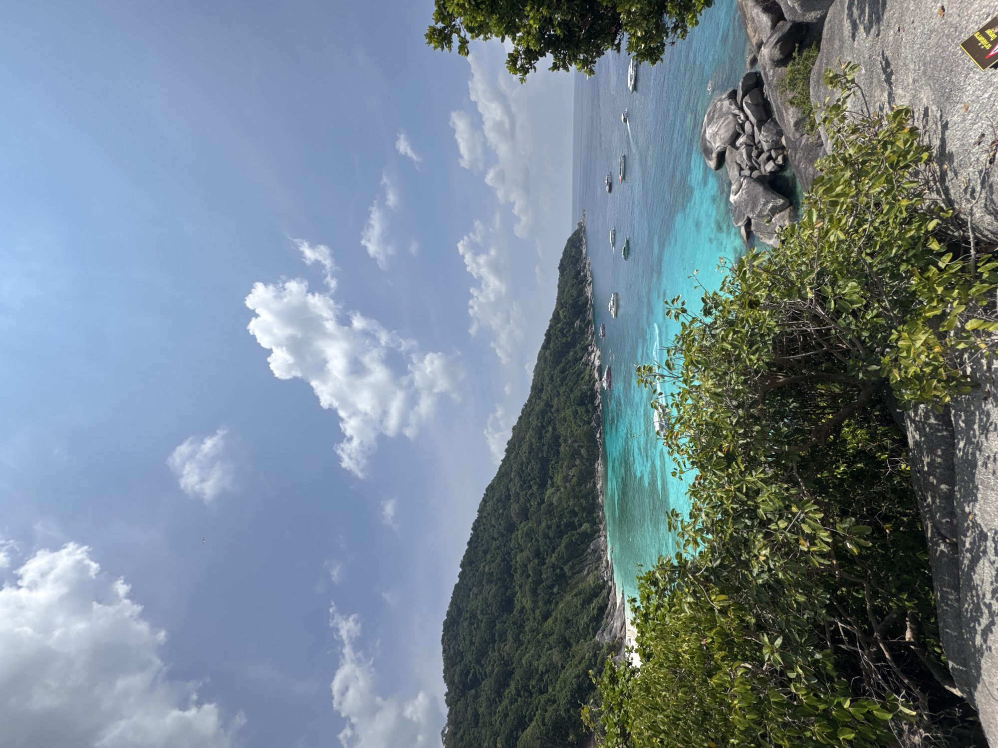 A vertical, outdoor shot depicts a tropical coastal scene likely from an elevated vantage point. The location is the Similan Islands, in Lam Kaen, Thailand. Azure water meets a rugged coastline, revealing numerous boats scattered along the shoreline. A lush, green hillside descends towards a narrow white sand beach. The upper part of the frame showcases a blue sky speckled with white, puffy clouds. In the foreground, leafy green foliage and rocky terrain obscure parts of the scenery. A sign is partially visible.