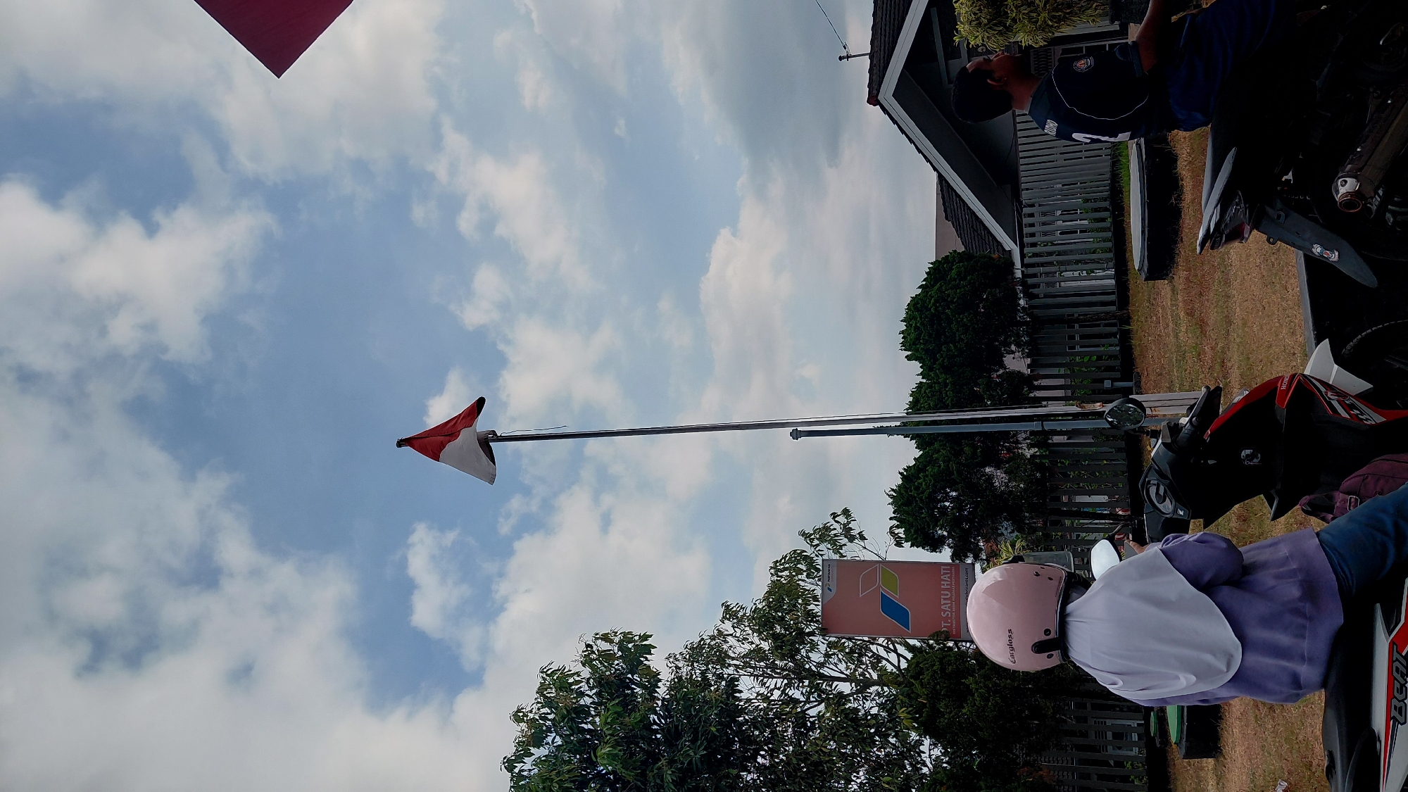 The image shows a flag pole with the Indonesian flag, which is red and white, flying against a cloudy blue sky. To the left of the flag pole, there is a tall tree and a vertical banner with the text "PT. SATU HATI". In the foreground, a person wearing a light purple hijab and a pink helmet is seated on a white and red motorcycle. The person is looking down. To the right of this motorcycle, another person in a dark blue shirt with the number "2" on the back is seated on a black motorcycle. They are looking to the right. The background features a fence, several trees, and a house with a dark roof. The ground is covered in dry grass and some dark circular objects. The scene appears to be outdoors in an urban or suburban setting, likely in the city of Rejasari, Indonesia, given the presence of the Indonesian flag and the banner text.