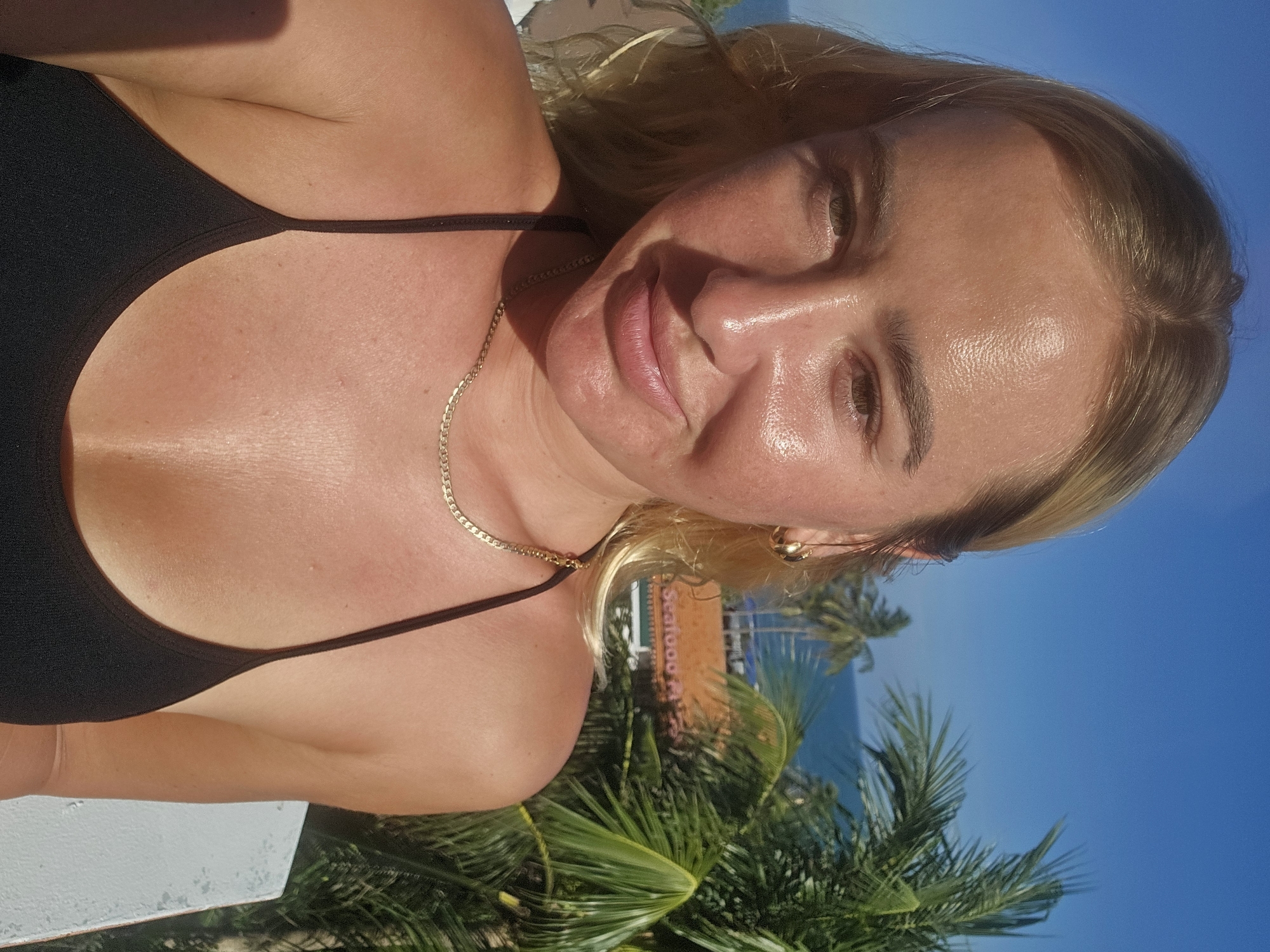 A blonde woman is photographed from the chest up, looking at the camera, with a slight smile. She is wearing a black bikini top and a gold chain necklace. Behind her, palm trees and a blue ocean are visible, suggesting a tropical coastal location. A building with a sign that appears to read "Seafood P..." can be seen in the background, partially obscured by palm leaves. The sunlight creates highlights on her skin, indicating a bright, outdoor setting. The overall impression is a candid snapshot taken on a sunny day in a seaside resort area.