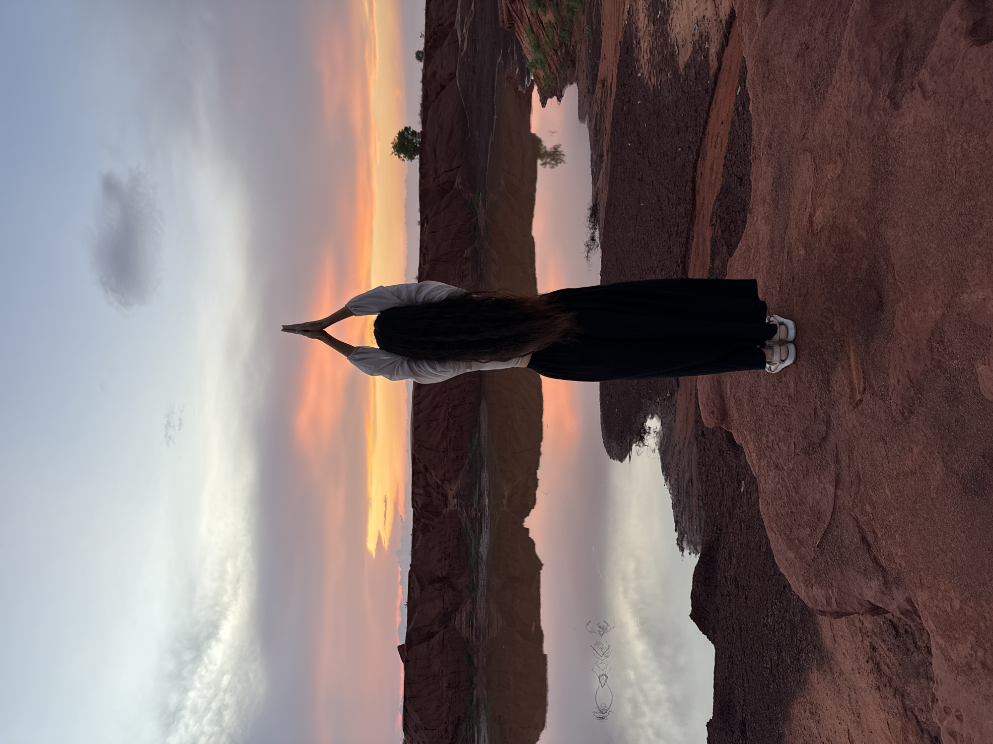 The image captures a serene natural landscape at either sunrise or sunset, featuring a person viewed from behind in Mui Ne, Vietnam. The individual has long dark hair and is wearing a white long-sleeved top and black wide-legged pants. Their arms are raised above their head, with hands pressed together in a prayer-like or meditative pose, creating a diamond shape against the sky.
The scene is dominated by striking red rock formations and cliffs, particularly prominent on the right side and in the background. A calm body of water occupies the middle ground, creating a near-perfect reflection of the sky and the surrounding landscape. The sky exhibits a beautiful transition of colors, from soft blue and grey on one side to vibrant oranges, pinks, and purples where the sun is setting or rising. Scattered clouds are visible, both in the actual sky and reflected on the still water. Small trees can be seen sparsely dotting the distant red land, their forms also mirrored in the tranquil water. The overall composition highlights the symmetry created by the reflection, fostering a sense of peace and contemplation. No visible text is present in the image.