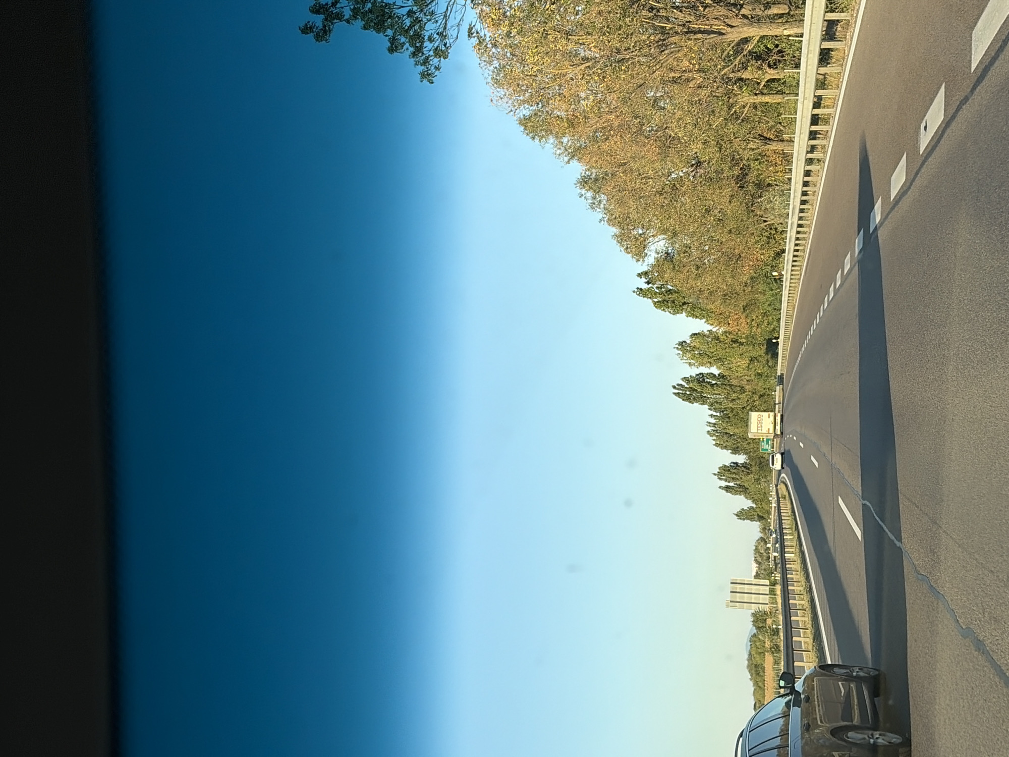 The image shows a view from inside a vehicle traveling on a highway. The road is paved, with white lane markings. On the left side of the road, there is a dark-colored SUV. On the right side of the road, there are trees with autumn foliage and a metal guardrail. In the distance, there are other vehicles, including a white car and a white truck with "TESCO" branding. A signpost is visible on the left side of the road. The sky is clear and blue, transitioning to a lighter shade towards the horizon. The scene appears to be set in the countryside, likely near the city of Báb, Slovakia, based on the context provided.