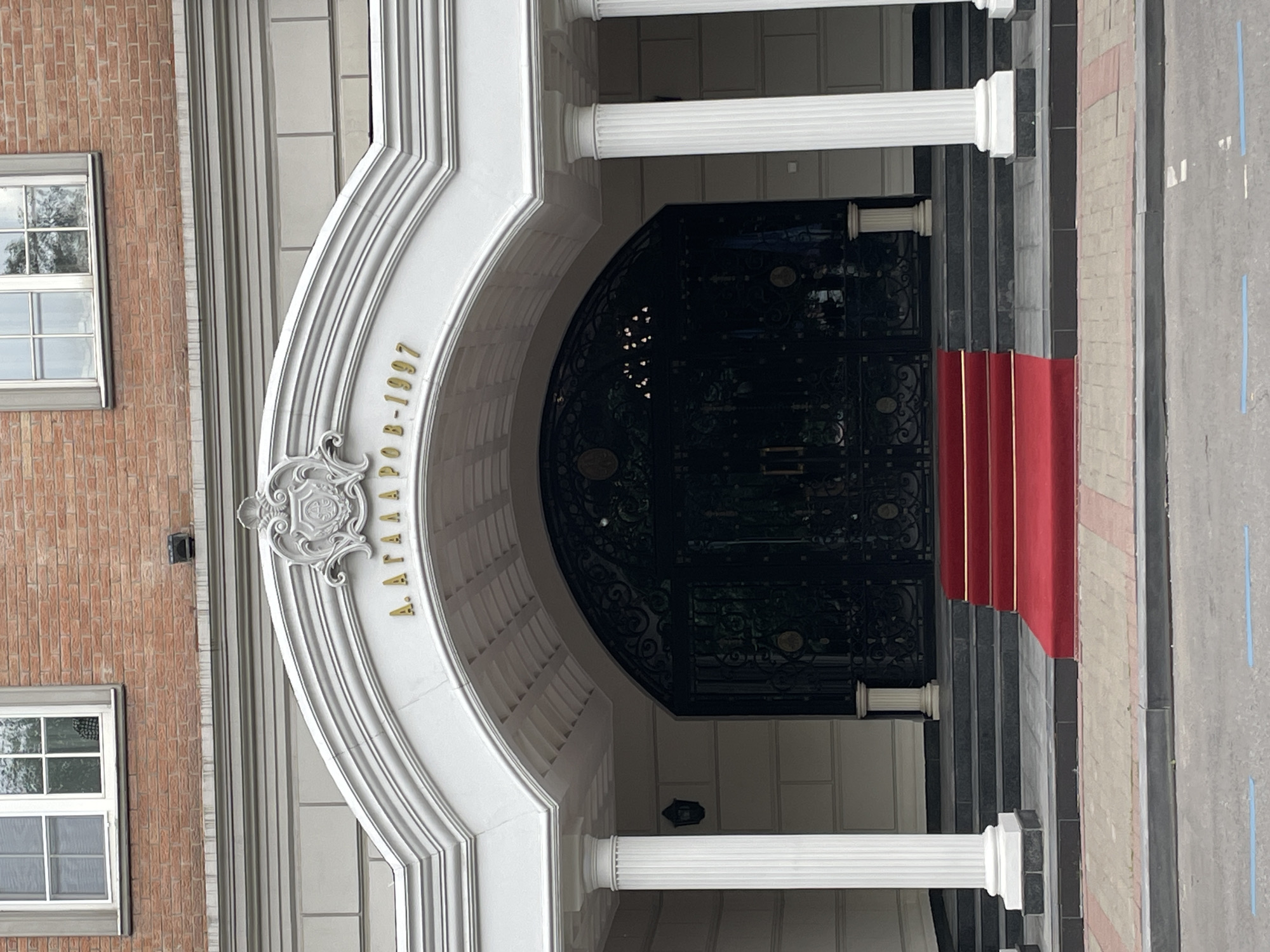 The image shows the entrance to a building, likely a private residence or business, located in Moscow, Russia. A red carpet with gold trim leads up a short flight of stairs to a grand, ornate doorway. Above the doorway, golden lettering reads "А. АГАБЯРОВ-1997". The entrance is flanked by tall, white fluted columns. The building's facade is made of brick with stone accents. The surrounding area appears to be a street with a sidewalk. There are no people visible in the image.