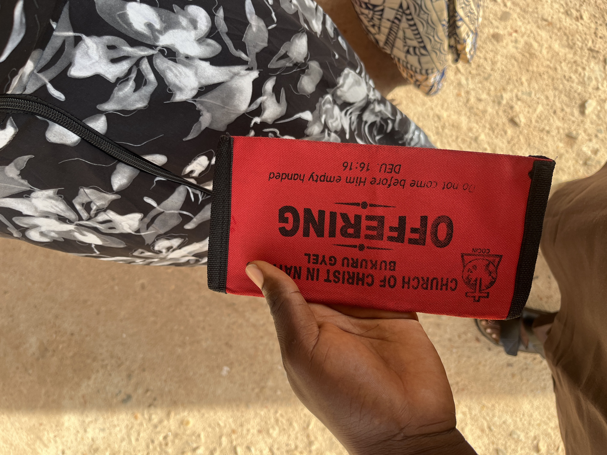 A close-up shot from a high angle shows a person's hand holding a red offering pouch. The pouch has black trim and features text in black lettering. The text on the pouch reads, "CHURCH OF CHRIST IN NIGERIA," "BUKURU GYEL," "OFFERING," and "Do not come before Him empty handed, DEU 16:16." Below "CHURCH OF CHRIST IN NIGERIA," there is a small logo that appears to be a rooster crowing above a cross. The background consists of a beige, sandy surface, and a black fabric with a white floral pattern. The person's feet, wearing dark sandals, are visible at the bottom of the frame, with brown fabric also visible. The overall impression is a detail of an offering pouch being held.