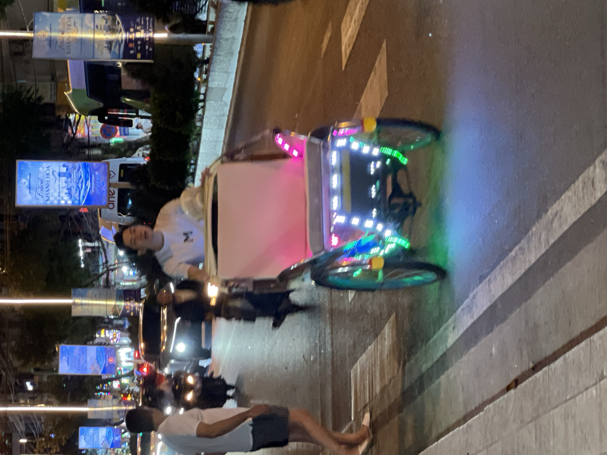 This is a nighttime street scene in Nha Trang, Vietnam, likely on a main road with traffic. In the foreground, a decorated cyclo, a three-wheeled bicycle taxi, is moving to the right. The cyclo is adorned with numerous colorful LED lights, predominantly pink and green, and a prominent yellow reflector. A person, possibly the driver, is seated in the front of the cyclo, wearing a white t-shirt with a letter 'M' printed on it. Behind the cyclo, a pedestrian with dark hair and a white t-shirt is walking away from the camera, to the left. In the background, several vehicles, including cars and motorcycles, are visible, their headlights and taillights illuminated. Overhead, tall light poles with hanging banners are present, one of which displays text that appears to be "KHANH HOA," a province in Vietnam. The street is paved and has visible pedestrian crossings. The overall atmosphere suggests a bustling urban environment at night.