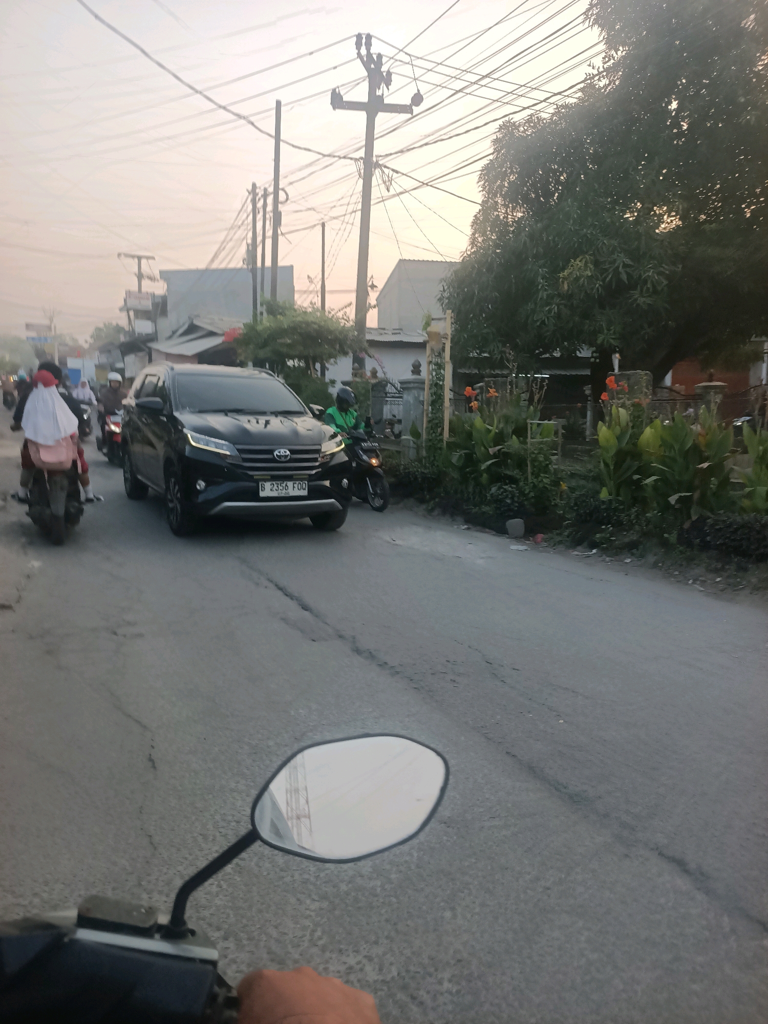The image depicts a street scene in Wanajaya, Indonesia, during what appears to be early morning or late afternoon given the soft light. The perspective is from the viewpoint of someone riding a motorcycle, with the handlebars and a rearview mirror visible in the foreground.
A black Toyota Rush SUV is the primary vehicle in focus, positioned in the center of the road and traveling in the same direction as the viewer. The SUV displays a license plate reading "B 2356 FOQ". Several motorcycles are also present in the scene. One motorcycle is to the left of the SUV, carrying two individuals, one of whom is wearing a white hijab and a pink backpack. Another motorcycle is to the right of the SUV, with a rider wearing a green jacket and helmet.
The environment features a paved road with some visible cracks and imperfections. Along the right side of the road, there is a dense arrangement of greenery, including large-leaved plants and flowers with orange blossoms, suggesting a residential or garden area. A tall tree with dense foliage stands behind these plants. In the background, there are buildings, some appearing to be single-story houses, and utility poles with a complex network of overhead electrical wires. The sky is a pale, hazy color, indicating either sunrise or sunset. The overall impression is of a busy, local street with typical urban and suburban elements.