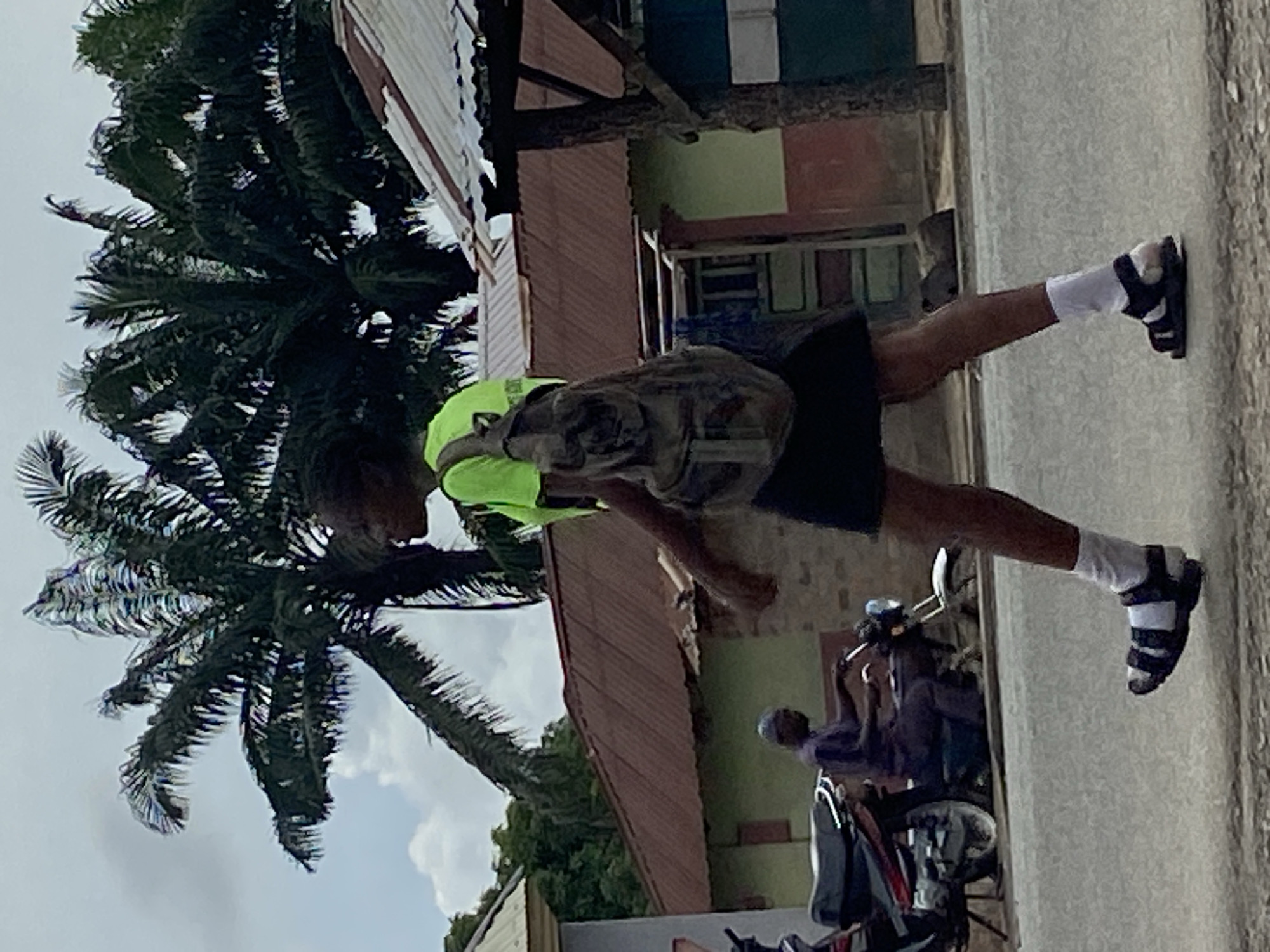 A street scene in Mararaba, Nigeria, shows a young person wearing a fluorescent yellow shirt, dark shorts, white socks, and black sandals walking on a sidewalk or paved area. The person is carrying a backpack and moving from left to right. A palm tree is visible on the left side of the image. To the right and behind the person are buildings constructed with corrugated metal and other materials, some appearing to be in disrepair. A parked motorcycle with a rider is observed along the sidewalk near the walking person. The sky appears overcast.