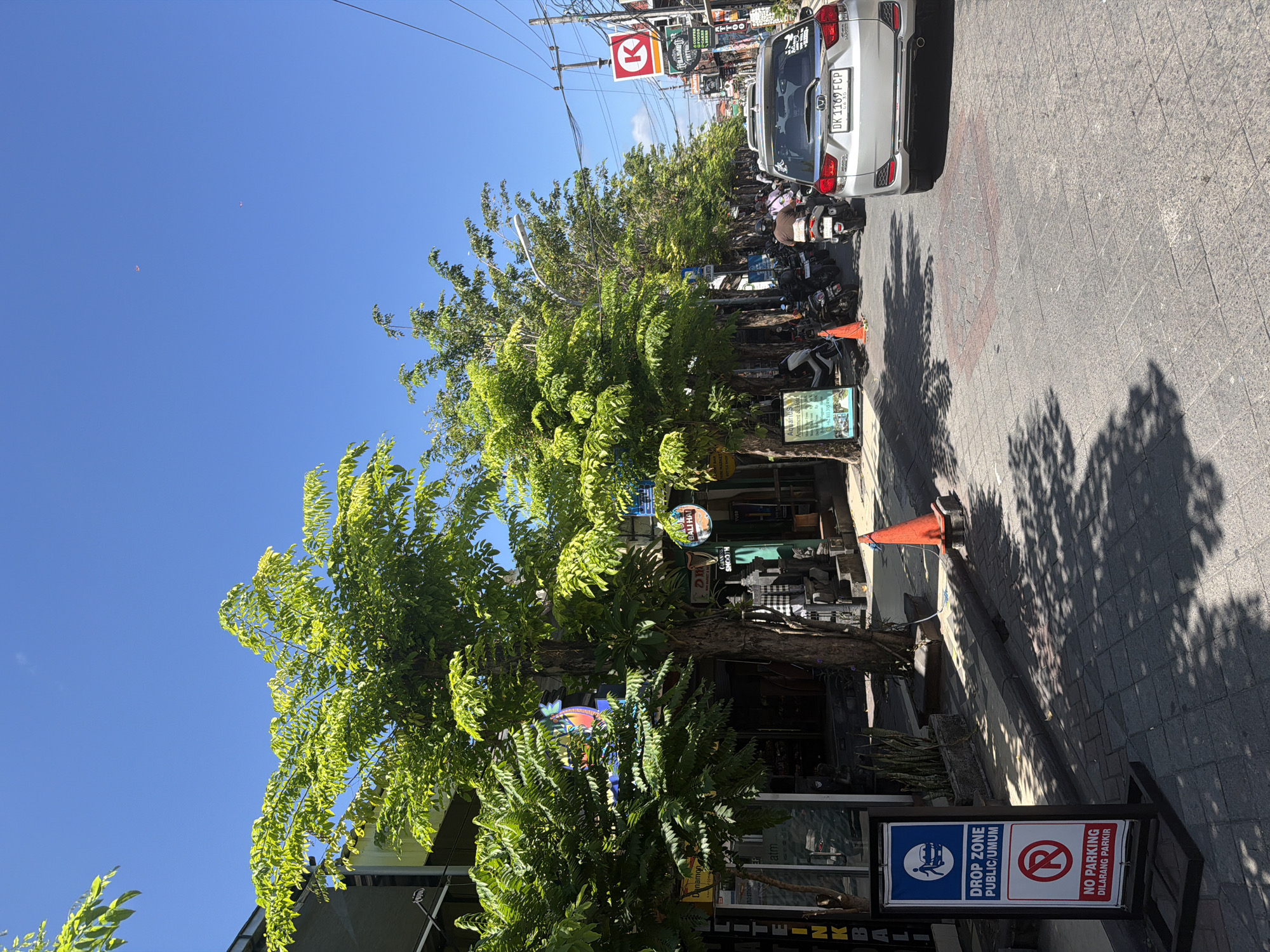 The image presents a sun-drenched street scene in Kuta, Indonesia, characterized by clear blue skies and strong shadows indicative of midday or early afternoon. An urban street is lined with numerous trees featuring vibrant green foliage, casting dappled patterns across the tiled sidewalk. Overhead, power lines crisscross the upper portion of the frame.
On the right side of the road, a silver SUV, identified by its license plate "DK 1169 FCP," is parked, followed by a cluster of motorcycles and scooters. A person in brown attire is visible near one of the parked scooters.
The street is flanked by commercial buildings, many with signs partially obscured by the trees and infrastructure. Visible business names and logos include "GUINNESS," "SMOOTH," "BALI HAI," and "ATTOO," along with a red and white "K" logo, and an "atm" sign.
In the foreground, a prominent black and white sign on a stand provides instructions and warnings. One panel designates a "DROP ZONE PUBLIC/UMUM," featuring an icon of a person with luggage, indicating an area for passenger pick-up or drop-off. The adjacent panel strictly warns "NO PARKING DILARANG PARKIR," accompanied by a red no-entry symbol. Two orange traffic cones are placed on the sidewalk near this sign. High in the sky, two tiny, distant objects, possibly kites, can be observed. The Indonesian text "UMUM" and "DILARANG PARKIR," along with the "DK" license plate prefix, confirm the location.