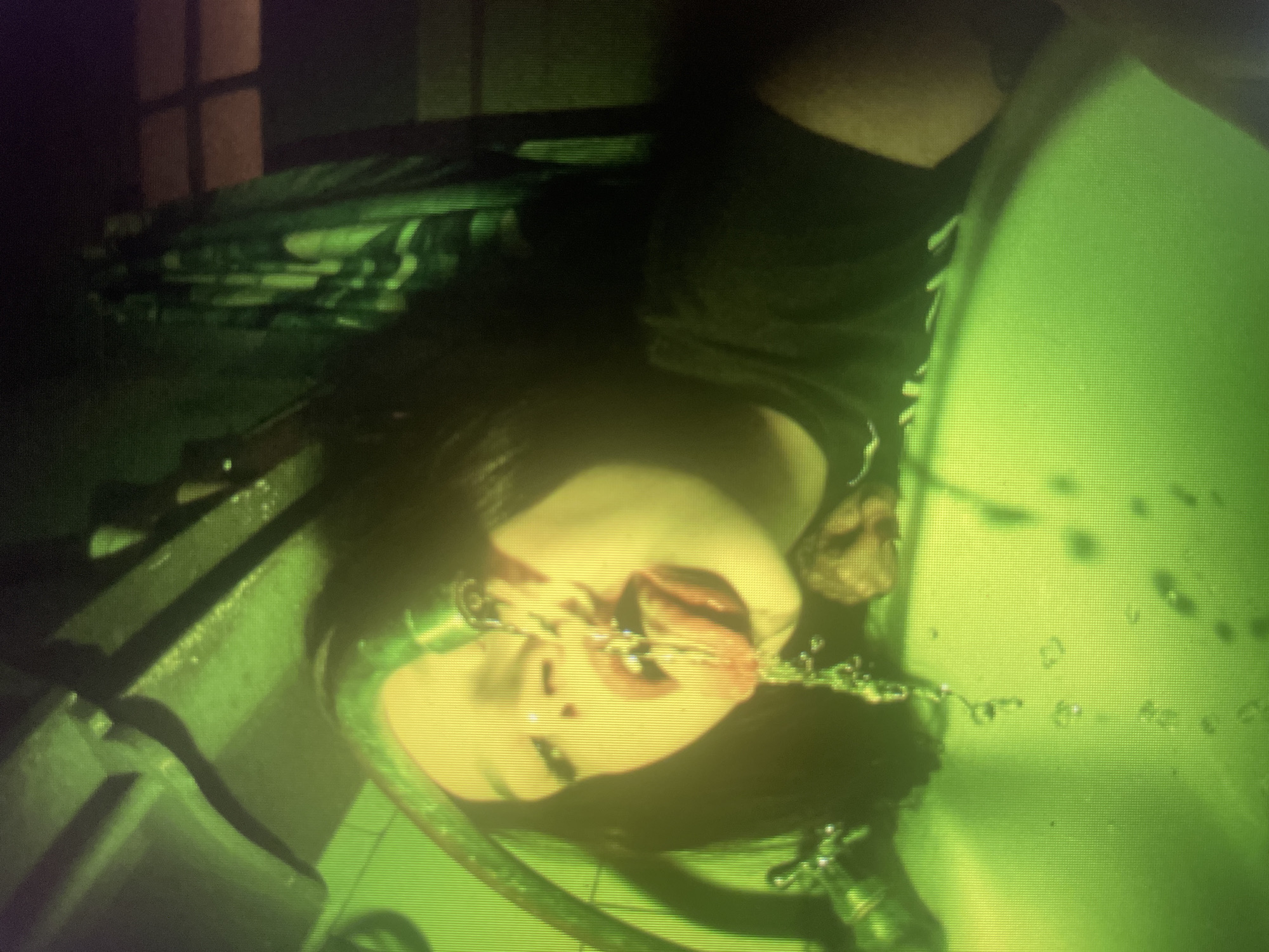 The image shows a close-up of a person drinking water from a faucet in a sink. The person's face is partially obscured by water, and their tongue is extended to catch the stream. The lighting is predominantly green, casting an unusual hue over the scene. The sink appears to be made of white porcelain, and there is a faucet with water running from it. The background is dark and indistinct, with some patterned textures visible, possibly a shower curtain or tiles. The overall impression is one of an intimate and perhaps unconventional moment. The location is not specified, but the presence of a sink and faucet suggests an indoor setting such as a bathroom or kitchen.