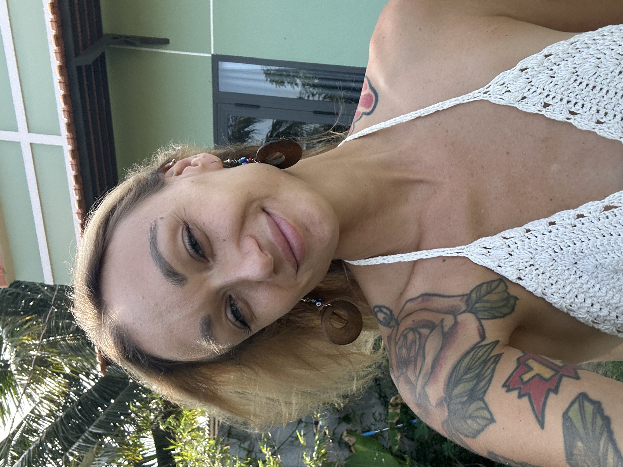 A medium-shot portrait depicts a fair-skinned woman with light blonde hair. She is smiling slightly, and her eyes are partially closed. She is wearing a white, crocheted top with thin straps. She is accessorized with circular wooden earrings, each featuring an "evil eye" bead. Her arm is visible, displaying a detailed rose tattoo, as well as another tattoo that includes red and yellow. The background consists of a light green building with dark-framed windows and a red-tiled roof. Palm trees and other greenery are visible in the lower left corner, bathed in sunlight, suggesting an outdoor setting in a tropical climate, most likely in Mũi Né, Vietnam.
