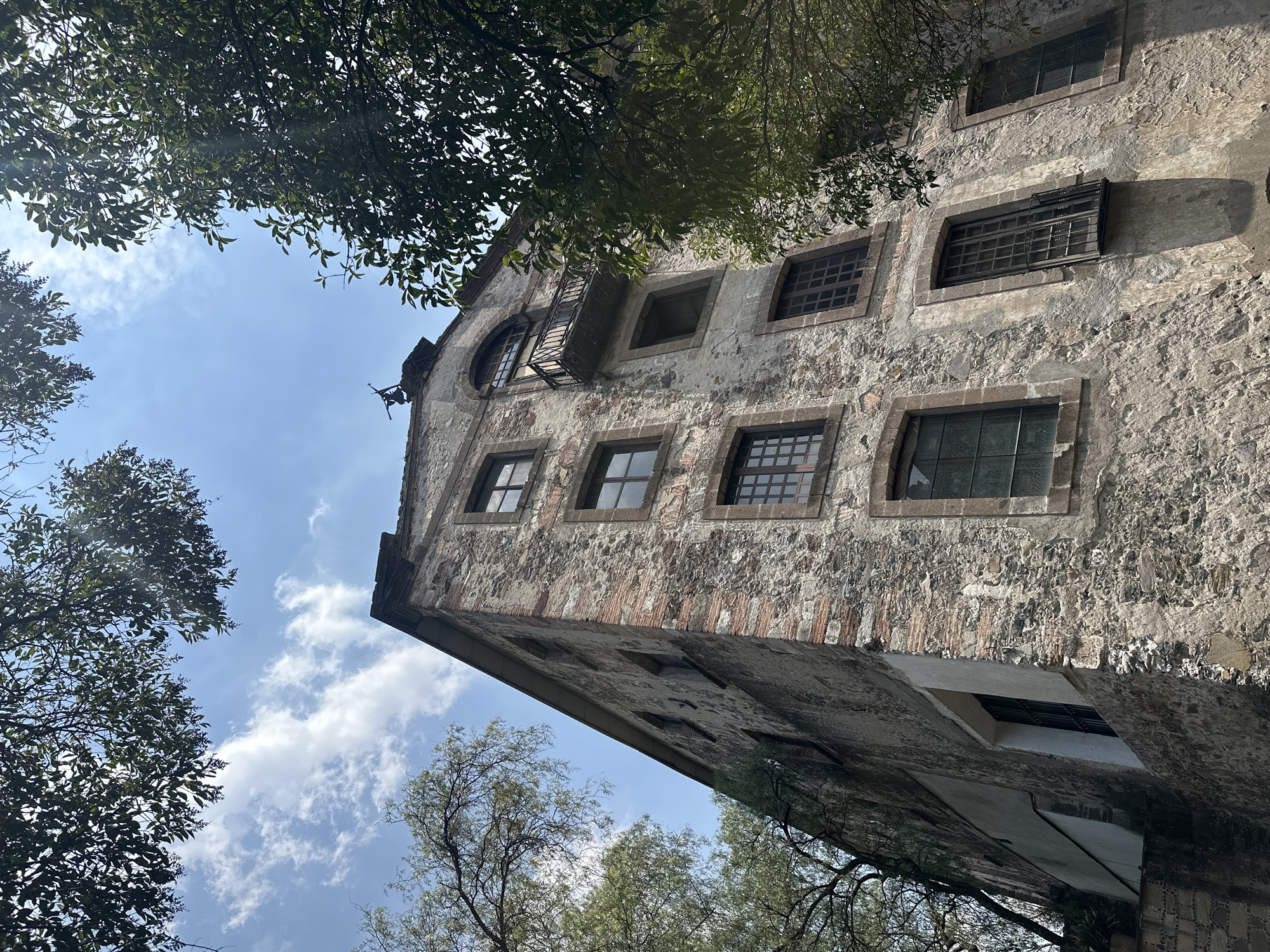 The image depicts a tall, old stone building with multiple windows, some with bars, framed by trees and the sky. The building appears to be deteriorating, with patches of discoloration and missing plaster. There are hints of brickwork beneath the stone facade. The image is taken from a low angle, looking up. Based on its architectural style and visible features, it is likely located in Tlalnepantla, Mexico, given its mention. The windows are rectangular, except for one arched window at the top. There is a small balcony or ledge near the top of the building. The sky is blue with scattered clouds. No people are visible.