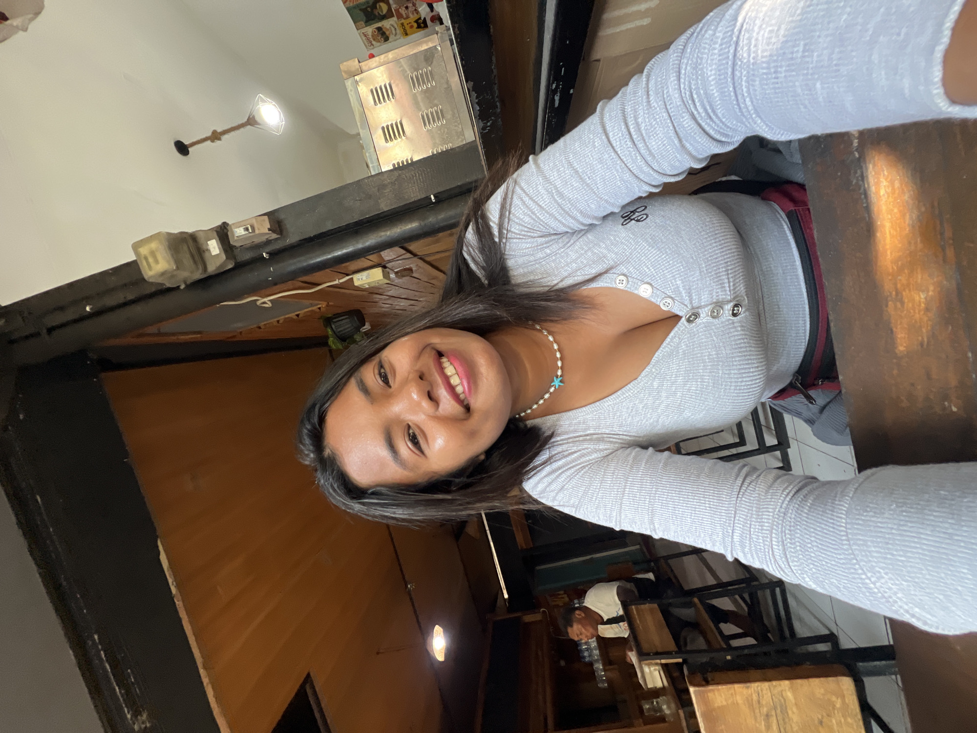 In Kuta, Indonesia, a young woman with dark hair smiles at the camera, her head slightly tilted. She is wearing a light gray, long-sleeved shirt with a pearl necklace featuring a turquoise pendant. The shirt has a V-neckline with three visible buttons. Behind her, a man wearing a white t-shirt sits at a wooden table in what appears to be an open-air restaurant or cafe. The background reveals wooden paneling on the ceiling and walls, a black metal pillar, and a metallic appliance on a counter. A light fixture hangs from the ceiling. Sunlight casts a warm glow on the scene.