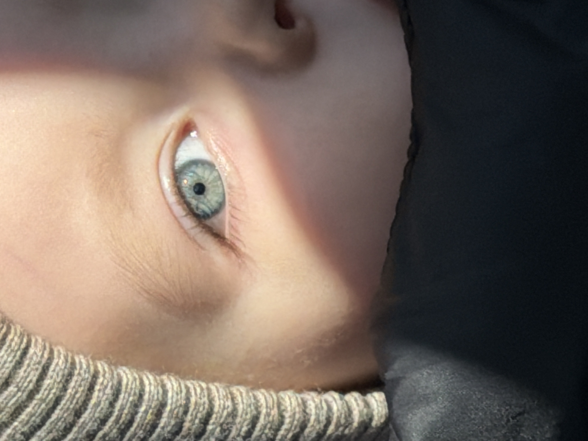A close-up shot of a child's face, specifically the eye and surrounding features. The child's eye is light blue with a visible pupil and eyelashes. The skin is pale and appears smooth. A portion of what seems to be a knitted sweater collar is visible at the bottom of the frame. To the right, there's a dark, undefined mass, potentially clothing. The image appears to be taken indoors with natural lighting casting a shadow across part of the face. There is no other information about the city of Chaikovsky, Russia.