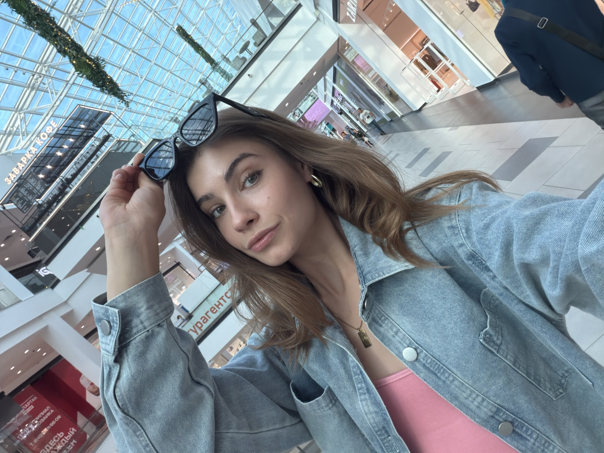 A young woman is taking a selfie inside a spacious, modern shopping mall located in Moscow, Russia. She has light brown hair, green eyes, and a neutral expression, looking directly at the camera. She is wearing a light blue denim jacket over a pink top, a golden rectangular pendant necklace, and golden hoop earrings. Her right hand holds a pair of black sunglasses, which are resting on top of her head.
The mall features a high, transparent glass ceiling supported by a metal framework, allowing ample natural light to illuminate the interior. Greenery in decorative hanging baskets is visible suspended from the ceiling structure. Multiple levels of the mall are visible, connected by escalators and framed by glass railings. The polished floor has a patterned tile design.
In the background, other shoppers are visible, and various store facades line the walkways. Visible text on signs includes "ЗАВАРКА КОФЕ" (ZAVARKA KOFE), translating to "Brewed Coffee," indicating a cafe. A partially visible "ZARA" sign identifies a clothing store. Other signs display "ТУРАГЕНТС" (TURAGENT), likely for a "Travel Agency," "ПАРТАК" (PARTAK), "ЗДЕСЬ КАЖДЫЙ ДЕНЬ" (ZDES KAZHDY DEN), meaning "Here every day," "ИГРЫ" (IGRY) meaning "Games," and "ТЦ ВЯЗЬМЫ" (TC VYAZMY), referring to a "Shopping Center Vyazmy." The activity is a casual selfie, capturing a moment within the bright, daytime mall environment.