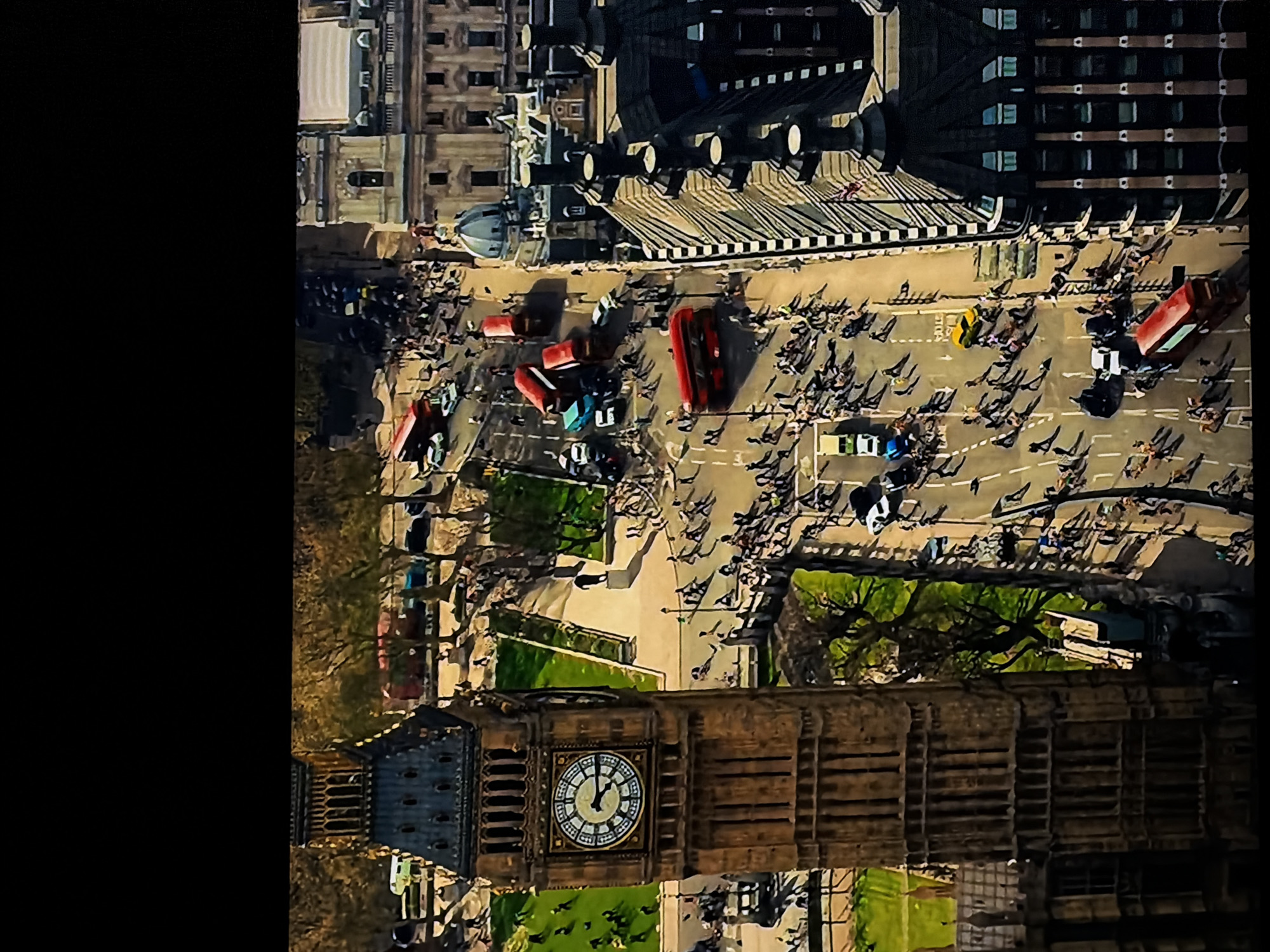 The image presents a high-angle or aerial view of a bustling urban scene, rotated 90 degrees counter-clockwise from its natural orientation. When correctly oriented, the iconic Elizabeth Tower (Big Ben) is prominently featured on the left, with its detailed clock face clearly showing the time as approximately 11:58.
The surrounding area is a busy street and pedestrian zone, populated by numerous people walking, indicating significant foot traffic. These pedestrians cast long, distinct shadows, suggesting a sunny day, likely in the late morning. Several vehicles are visible on the roads, including multiple distinctive red double-decker buses and a few yellow taxis.
In the background, there are large, ornate buildings with classical architecture, consistent with the Houses of Parliament complex. Interspersed throughout the scene are green spaces with trees. The combination of Big Ben, red double-decker buses, and yellow taxis strongly indicates the location as London, England. The activity captured is typical urban movement, showing the daily life and flow of people and traffic in a major city center. Aside from the Roman numerals on the clock face, no other legible text is visible in the image.