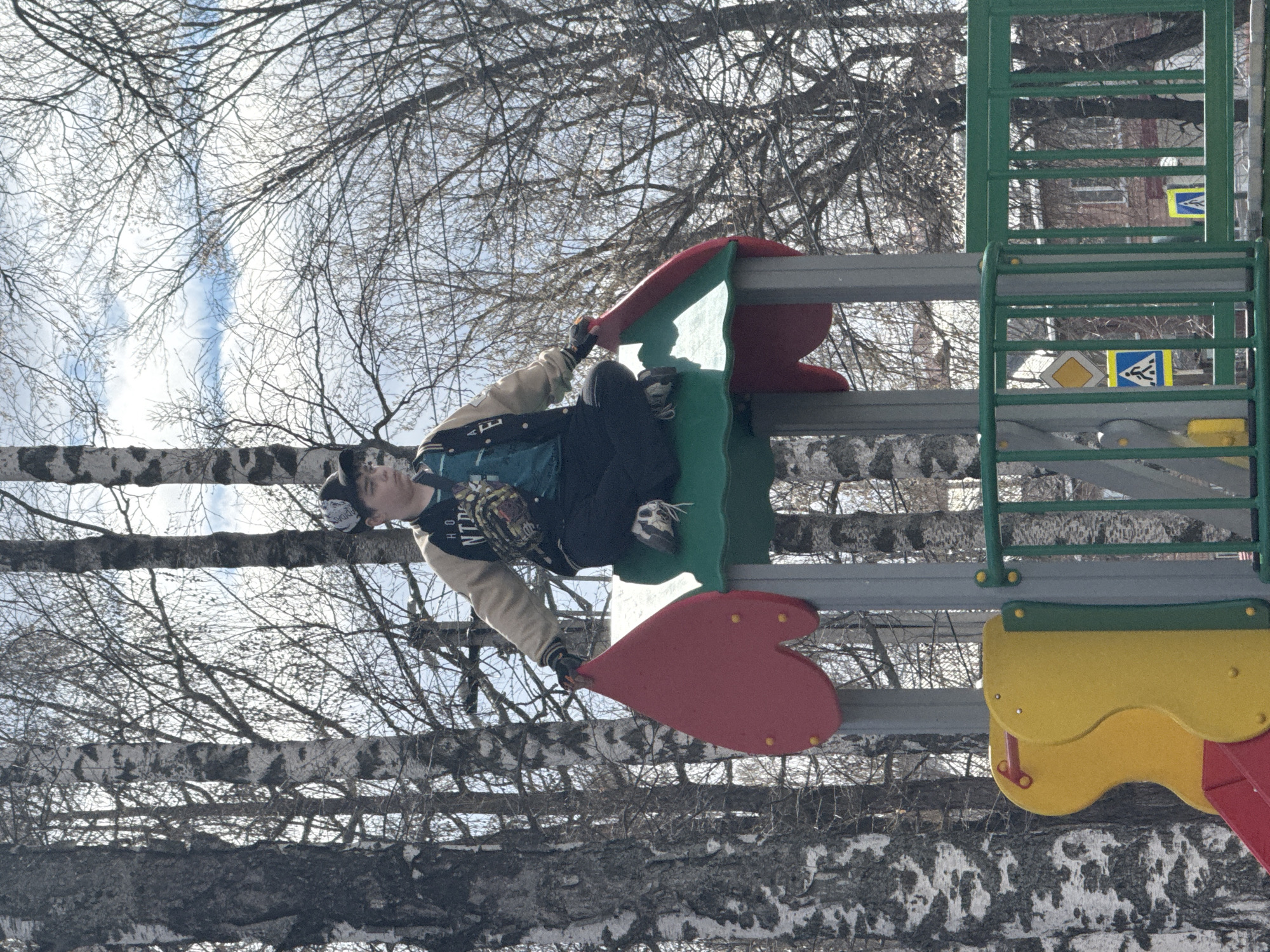 A young individual is positioned on a children's playground structure in what is likely the city of Zav'yalovo, Russia, as suggested by the context. The individual is wearing dark trousers, a light-colored jacket, a black t-shirt with graphics, and sneakers. He is holding onto the playground equipment. The playground structure itself consists of multiple components, including a slide in yellow and red, green ladders, and red heart-shaped elements. The environment includes birch trees, bare of leaves, and what appears to be a light, overcast sky. In the background, faint outlines of buildings and street signs are visible through the branches.