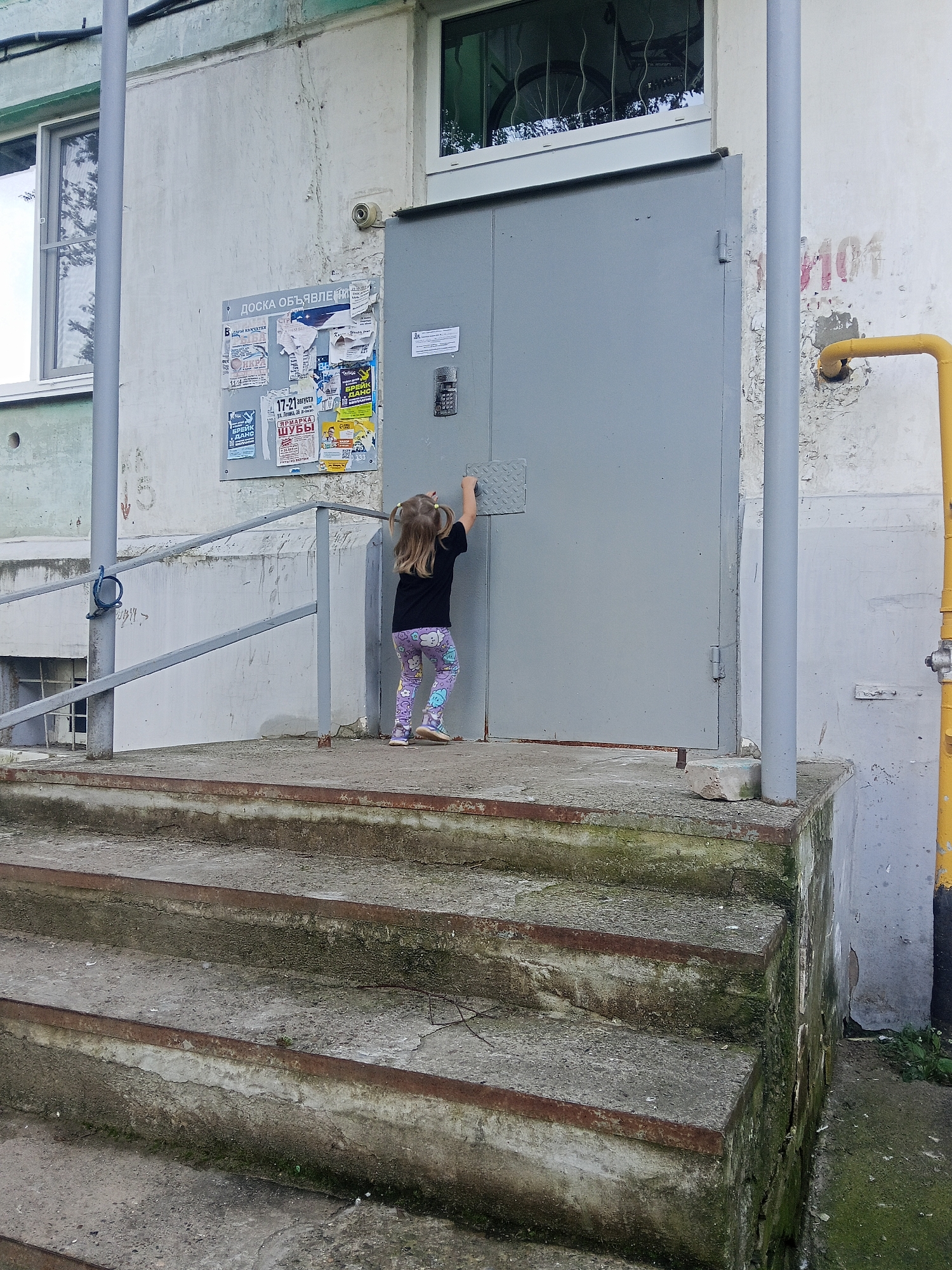 The image depicts a child's hand reaching for a metal door handle in an urban setting. The child, a young girl with blonde pigtails, is wearing a black t-shirt and colorful leggings. She is standing on a concrete landing in front of a large gray metal door. Adjacent to the door is a bulletin board covered in various posters and flyers, including some with Russian text. The building facade is concrete and shows signs of wear, with peeling paint and a surveillance camera mounted on the wall. A yellow pipe runs vertically along the side of the building. In the foreground, a set of weathered concrete stairs with rusted metal nosings leads up to the landing. The overall environment suggests a common residential building entrance in Chaikovsky, Russia.