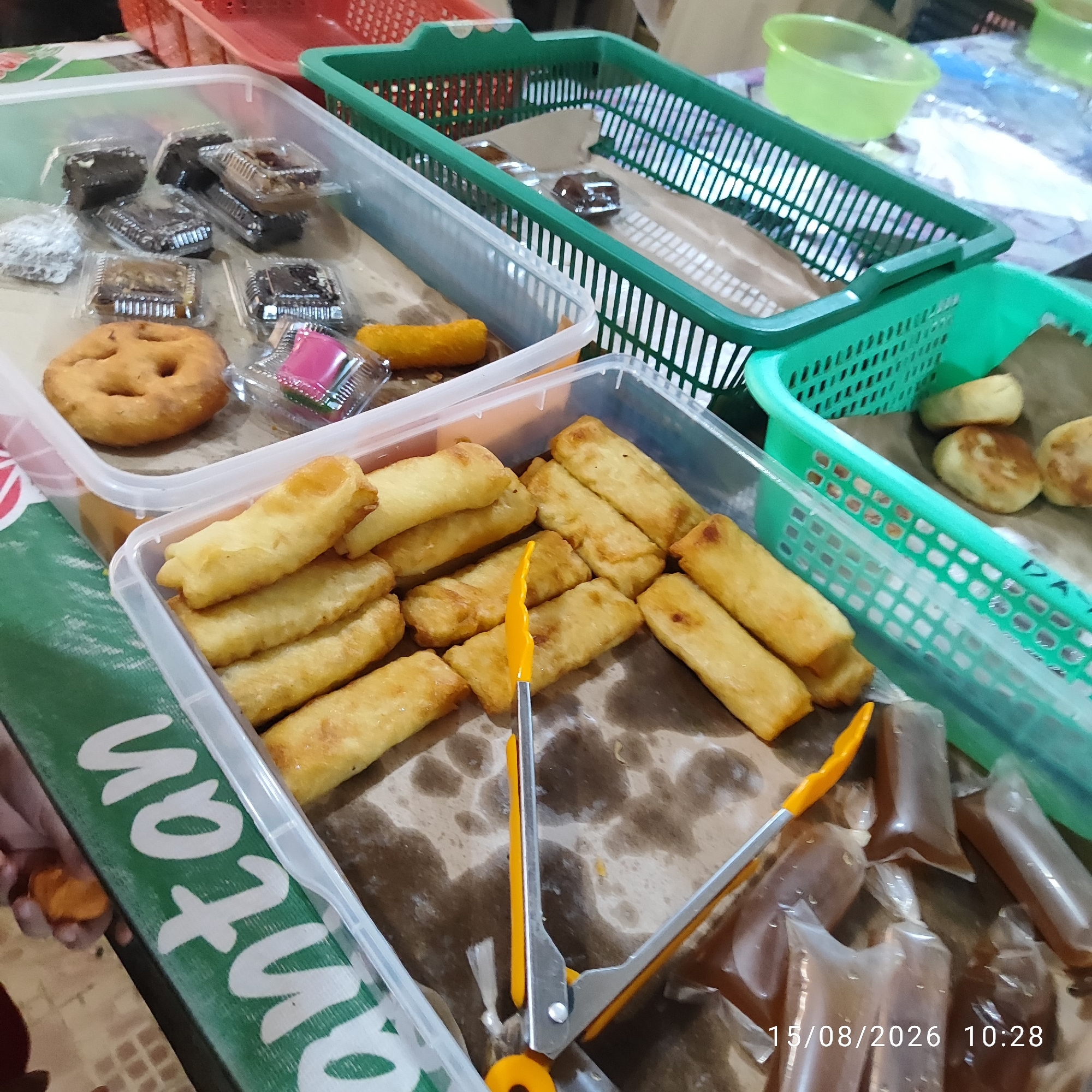 This image depicts a marketplace stall in Pembaharuan, Indonesia, showcasing a variety of Indonesian snacks and beverages. In the foreground, a clear plastic container holds numerous fried spring rolls, some of which are being picked up with yellow-handled tongs. To the left of the spring rolls, a single fried dough pastry with a smiley face design is visible. Behind this, another clear plastic container holds several small, individually packaged chocolate brownies and some other unidentified snacks. To the right of the spring rolls, a clear plastic bag contains what appears to be brown liquid, possibly a beverage. In the background, green plastic baskets are stacked, containing more snacks. The stall appears to be set up on a table with a green cloth that has white lettering, partially obscured. The overall impression is a vibrant and bustling food stall, typical of Indonesian markets.