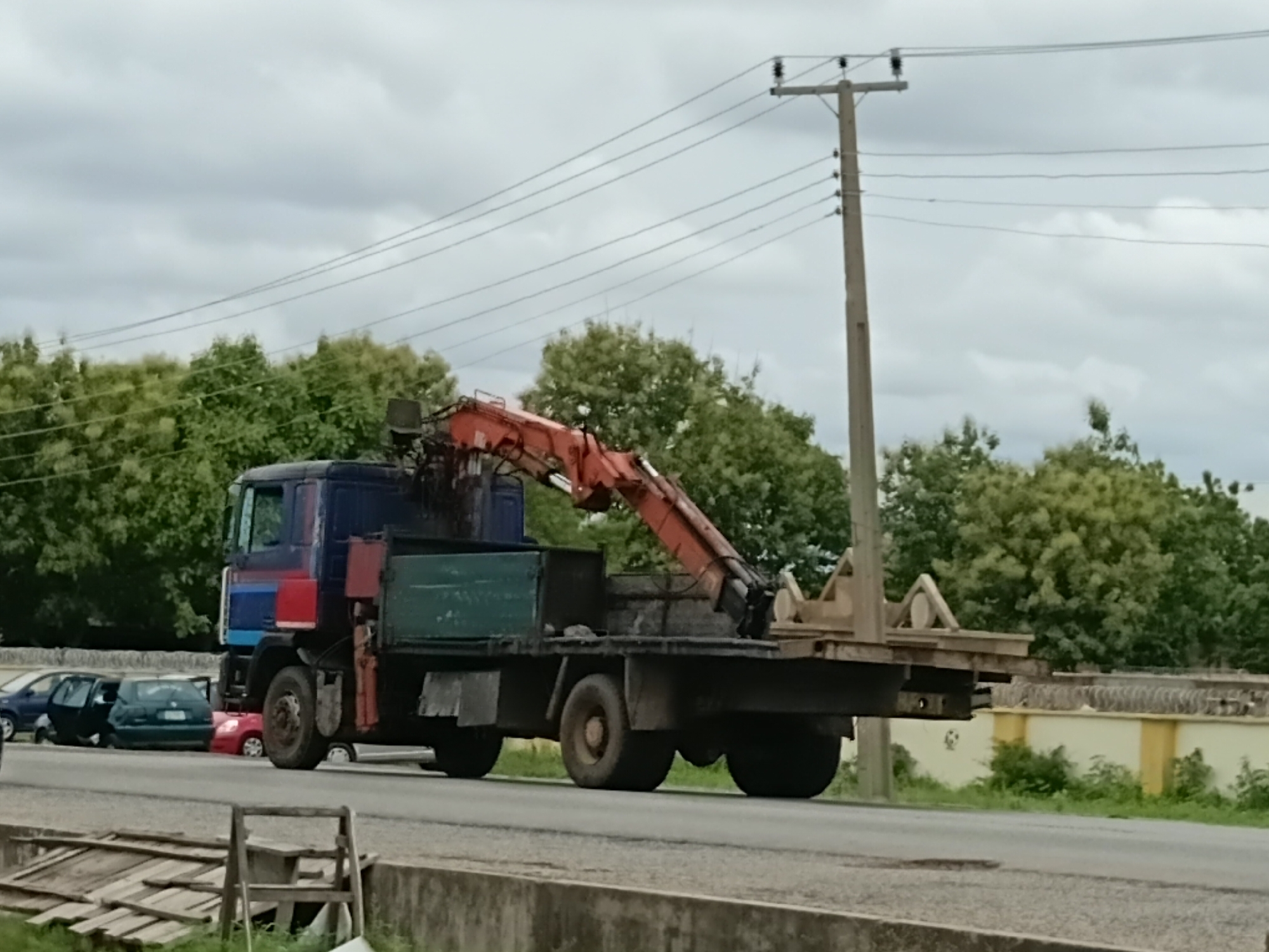 A blue and red truck with a crane attachment is parked on the side of a road in Jalingo, Nigeria. The truck is facing right, and the crane is extended. Several cars are parked behind the truck, and trees and a utility pole are visible in the background. The sky is cloudy. The scene appears to be daytime.