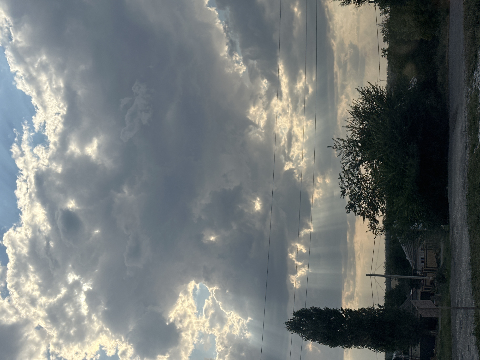 The image depicts a dramatic sky with crepuscular rays piercing through thick, dark clouds. Sunlight illuminates patches of the sky, creating a stark contrast. Below the sky, a tall, dark evergreen tree stands to the left, with a dense bush of green foliage to the right. In the background, several low-lying buildings with peaked roofs are visible, partially obscured by trees and vegetation. Utility poles and wires are present across the frame, indicating human infrastructure. The ground in the foreground appears to be a rough, gravelly surface. The scene is consistent with the outskirts of Kamenolomni, Russia, given the presence of rural architecture and natural elements.