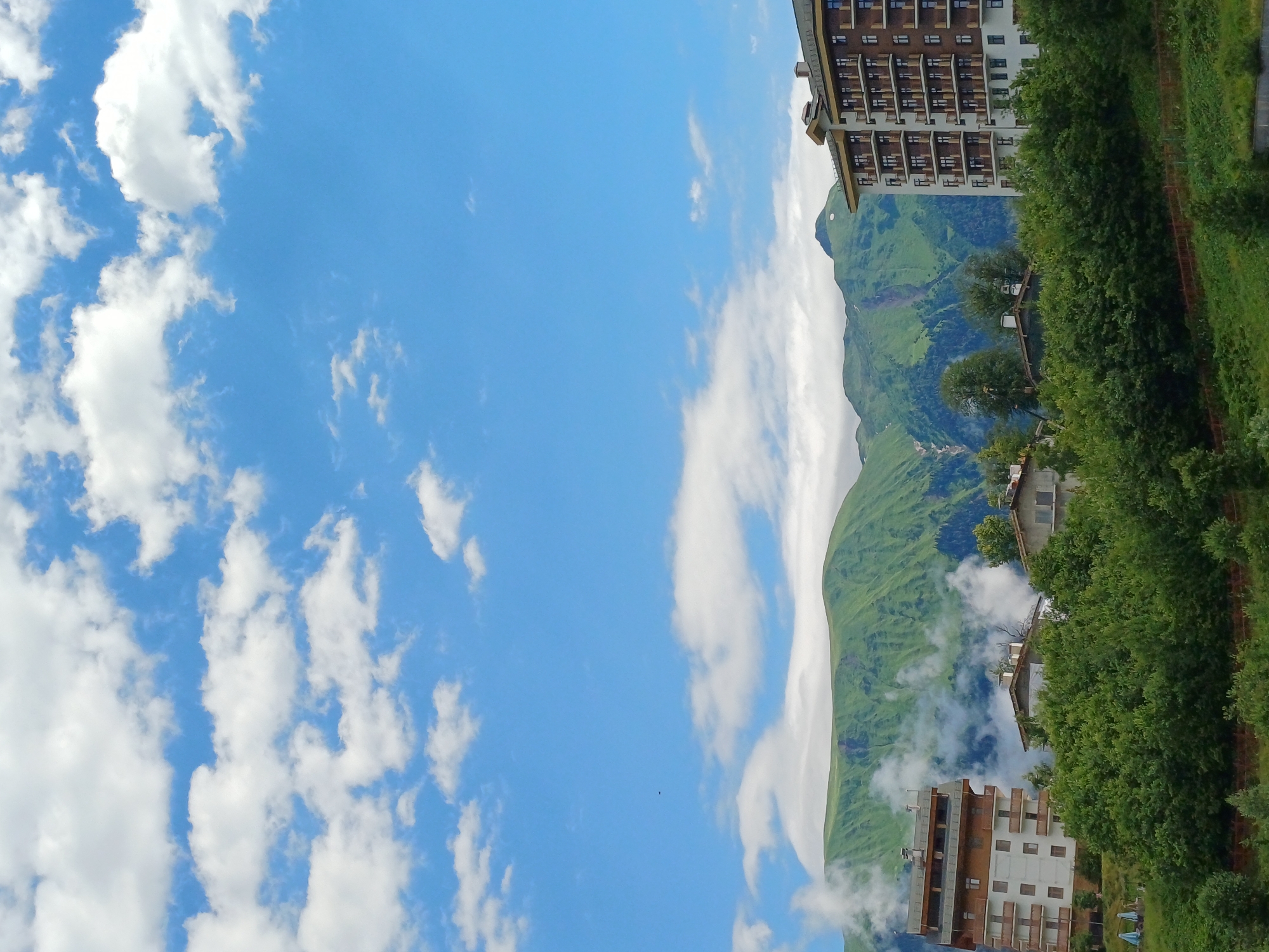 The image depicts a scenic mountain landscape with several buildings and lush greenery. In the foreground, dense green trees and shrubs are visible, with a subtle incline in the terrain. Several modern buildings are nestled among the trees. On the left side, a light-colored building with multiple balconies and a brown roof stands prominently. To its right, a cluster of smaller buildings with tiled roofs can be seen. On the far right, a taller, multi-story building with a brown facade and numerous balconies is present.
In the midground, rolling green mountains dominate the scene, with some areas showing patches of lighter green and possibly snow or rock. Wisps of white clouds or mist are clinging to the slopes of these mountains, adding a sense of depth and atmosphere.
The sky above is a vibrant blue, dotted with fluffy white cumulus clouds, indicating fair weather. A tiny dark speck, possibly a distant bird or aircraft, is visible in the upper left quadrant of the sky.
The location is identified as the city of Estosadok, Russia. The overall impression is one of a modern resort or residential area integrated into a natural mountain environment. There are no people or animals visible in the image. The scene is static and peaceful.