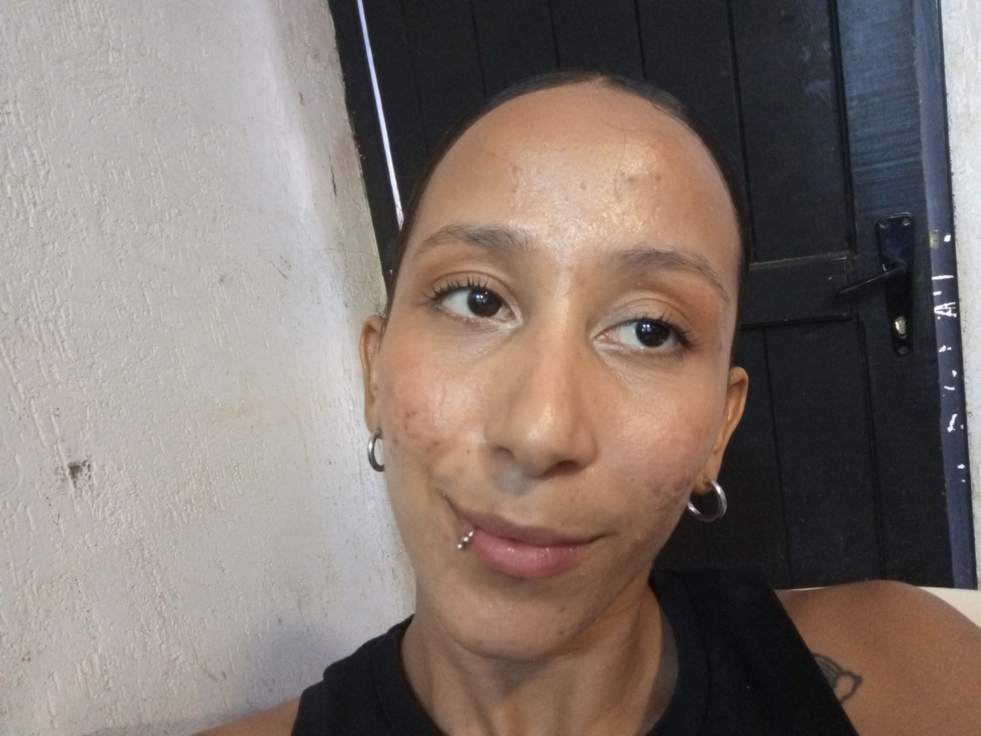A medium close-up portrait features a person with light brown skin, brown eyes, and no visible hair. The person has a lip piercing on the lower left side and hoop earrings. They are wearing a black tank top.
The background includes a textured white wall on the left and a dark gray or black door on the right. The door has a silver handle.
No specific details indicate the location is Porlamar, Venezuela.