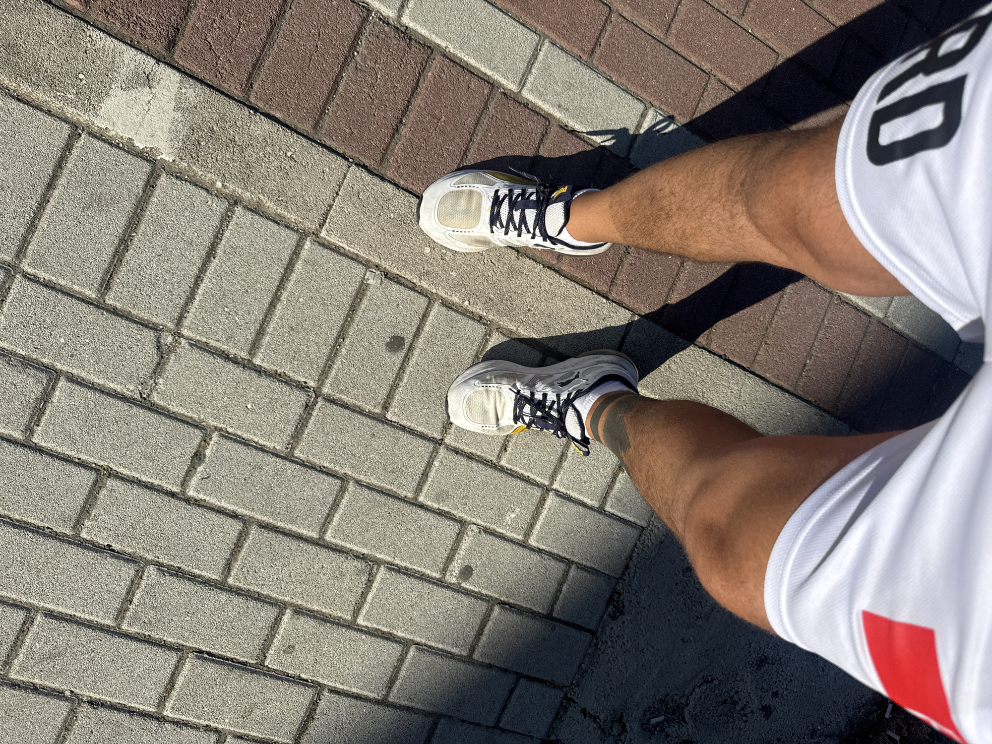 A top-down perspective captures the lower half of a person standing on a paved surface in Rafailovići, Montenegro. The ground consists of interlocking pavers; light gray on the left, transitioning to reddish-brown on the right, separated by a worn, lighter strip. A large, dark shadow covers the lower-left section of the gray pavers. The person's legs are tanned, muscular, and hairy. The left ankle displays a solid black band tattoo. Both feet wear white athletic sneakers with thick soles, dark laces, white ankle socks, and yellow detailing near the lacing. The person wears white athletic shorts; the left side features a red rectangular emblem, and the right side shows partial dark lettering, specifically "RD". The scene is brightly lit by strong sunlight, casting distinct shadows.