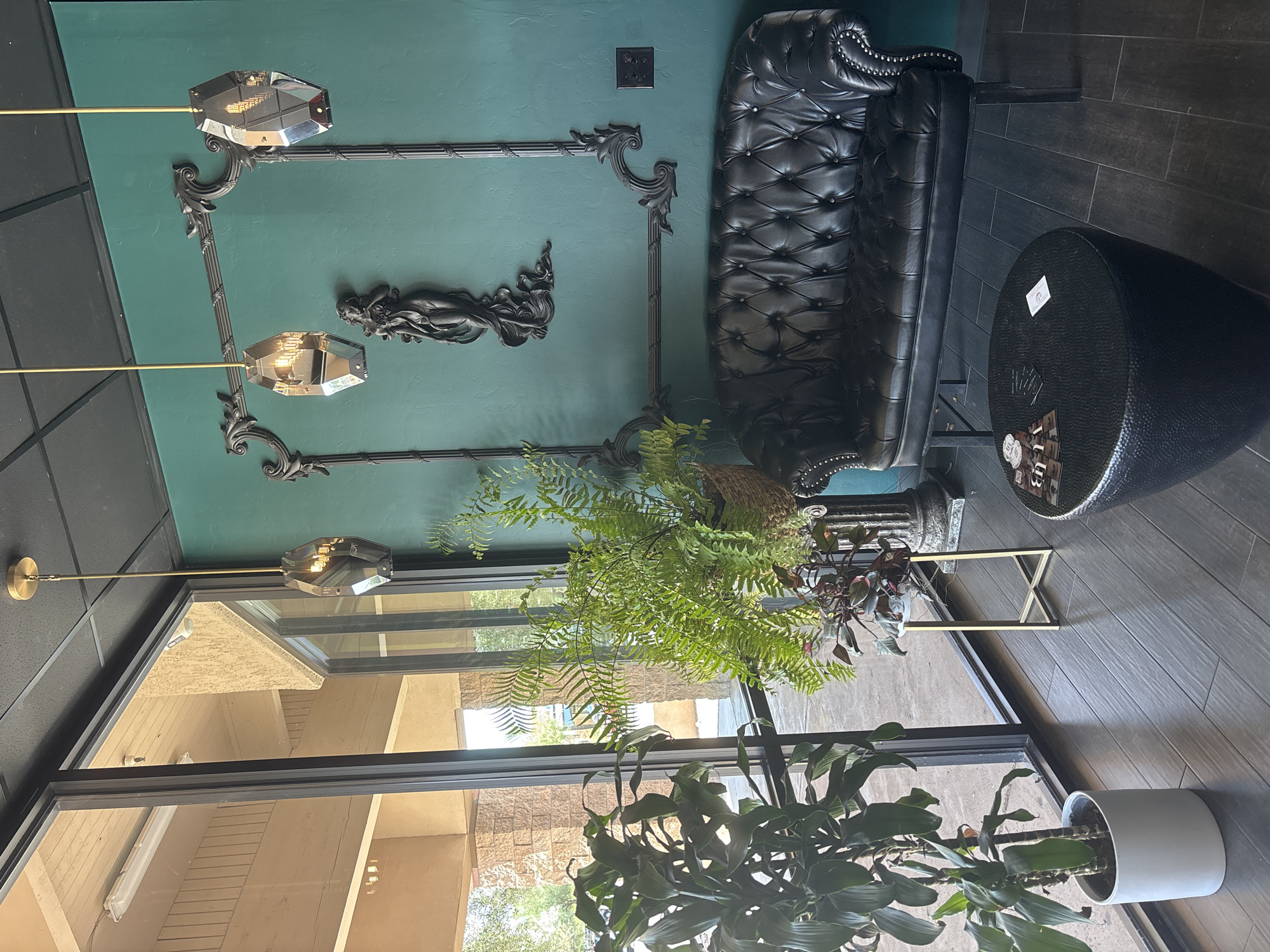 This is a wide shot of an indoor waiting area. The room is lit by natural light coming in from large windows on the left and also from three modern, geometric pendant lights hanging from the ceiling. The walls are painted a deep teal color and are decorated with ornate dark gray molding that forms a large, abstract rectangular frame. Inside the frame, a dark gray sculpture of a woman is mounted on the wall.
In the center of the room, a black tufted leather sofa sits against the teal wall. In front of the sofa is a round, textured black ottoman. On the ottoman are a few business cards and a small stack of pamphlets. To the left of the sofa, near the windows, are two potted plants. The larger plant on the left is a tall green plant with broad leaves in a white pot. To its right is a smaller fern in a woven basket. The floor is made of dark wood planks.
The exterior visible through the windows suggests a dry, sunny climate with adobe-style buildings and some sparse vegetation. The scene appears to be a modern and stylish waiting area, possibly for a business or salon.