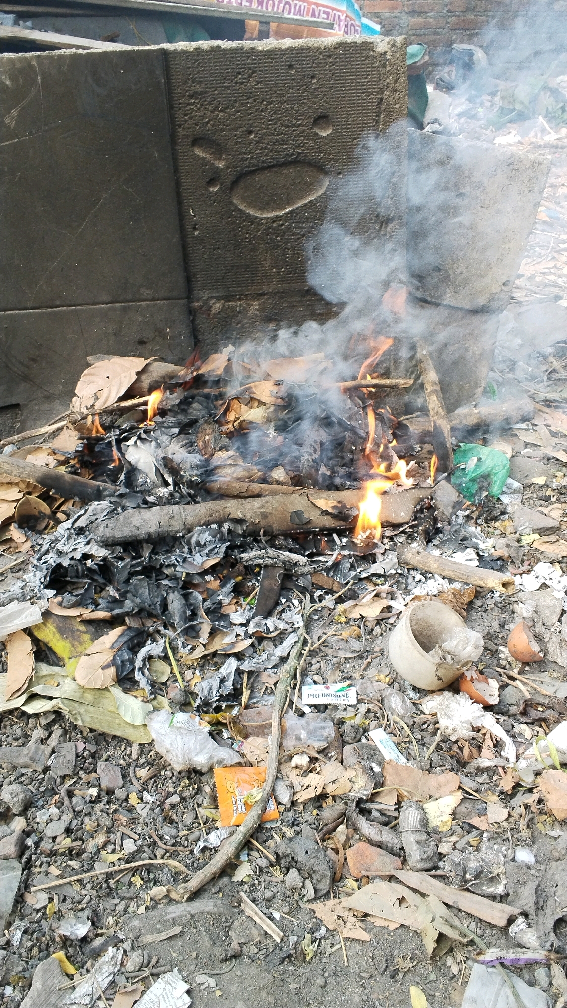 A small fire burns in a pile of dry leaves, twigs, and debris on the ground. Smoke rises from the fire, obscuring the background. The surrounding area is littered with more leaves, ash, and various pieces of trash, including plastic bags and a broken eggshell. In the background, there are two large, rough-textured concrete blocks. One block, on the left, is dark gray and smooth on its left side, with a textured surface on the right featuring an oval shape and several smaller dots. The other concrete block, on the right, is lighter in color and appears to be a similar rough texture. A blue and white sign with text is partially visible at the top of the image, behind the concrete blocks. The scene is outdoors, and the ground appears to be dirt and gravel. The overall impression is one of an outdoor waste disposal area, possibly in an urban or semi-urban setting. Based on the provided information, the location is Penompo, Indonesia.