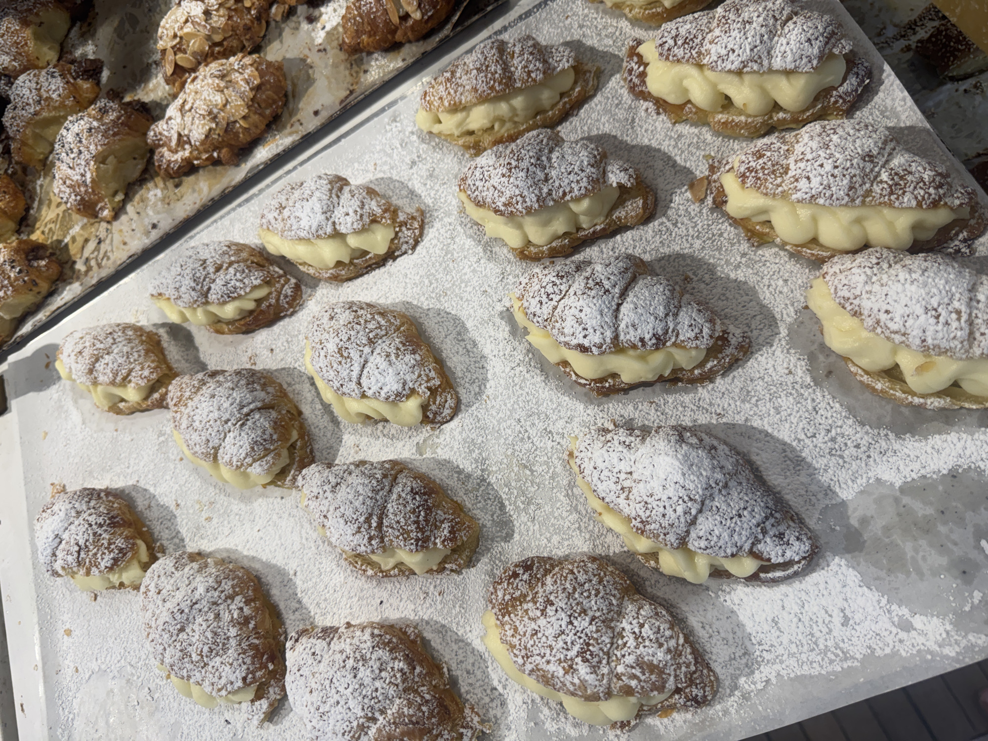 The image shows a tray of pastries, possibly in a bakery or shop in Ramat Gan, Israel. The pastries appear to be croissants, some plain and some filled with a creamy filling. All are heavily dusted with powdered sugar. There are no people visible in the image. The pastries are arranged on a light-colored surface, which could be a metal tray. The scene suggests food preparation or display for sale.