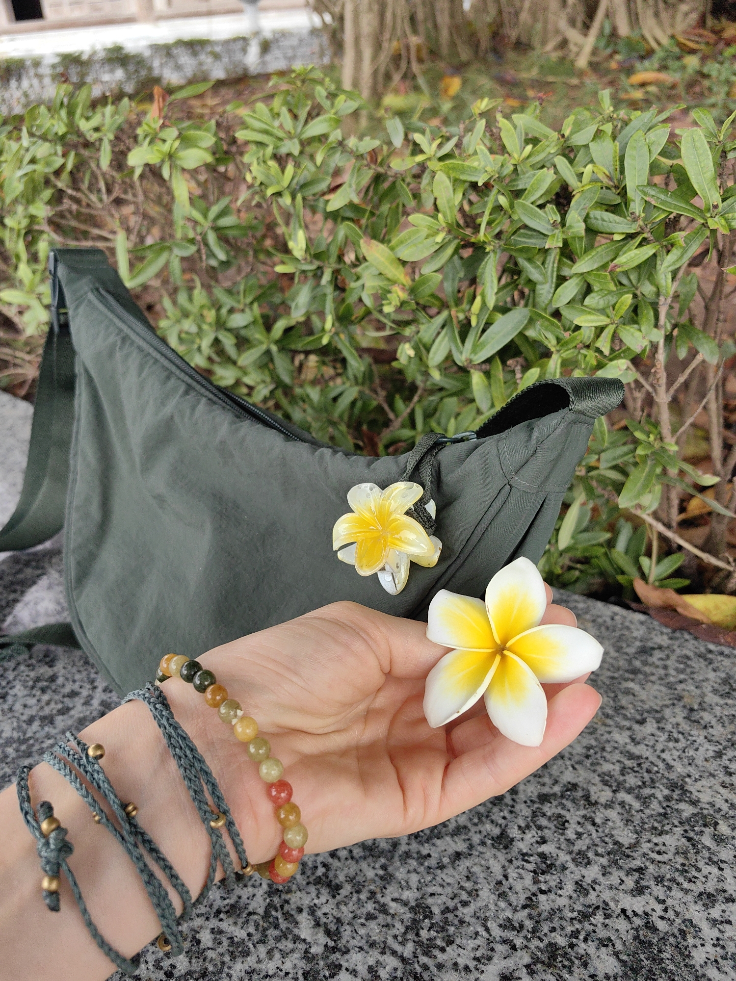 The image captures a close-up outdoor scene featuring a hand, a bag, and frangipani flowers, set against a backdrop of lush greenery and a stone surface in what is contextually Hong Gai Ward, Vietnam.
In the foreground, a light-skinned hand is central, gently holding a vibrant, real frangipani flower with white petals and a yellow center. The hand is adorned with two distinct bracelets. One is a beaded bracelet composed of spherical stones in a gradient of earthy colors, including green, yellow, orange, and reddish-brown. The second bracelet is a multi-strand, braided cord in a muted green-blue hue, featuring small metallic gold-colored beads and an adjustable knot design.
Adjacent to the hand, resting on a speckled gray and black granite-like surface, is a dark olive-green sling or crossbody bag. The bag appears to be made of a soft, lightweight fabric, possibly nylon, with visible seams and a black zipper closure. Attached to the zipper pull is an artificial, yellowish frangipani flower charm, echoing the real flower held in the hand.
The background is dominated by dense, healthy green foliage, suggesting well-maintained bushes or hedges typical of a garden or park setting. The natural, soft daylight indicates the photograph was taken during the day. The scene evokes a calm, pleasant atmosphere, showcasing a blend of natural beauty and personal accessories. There is no visible text in the image.