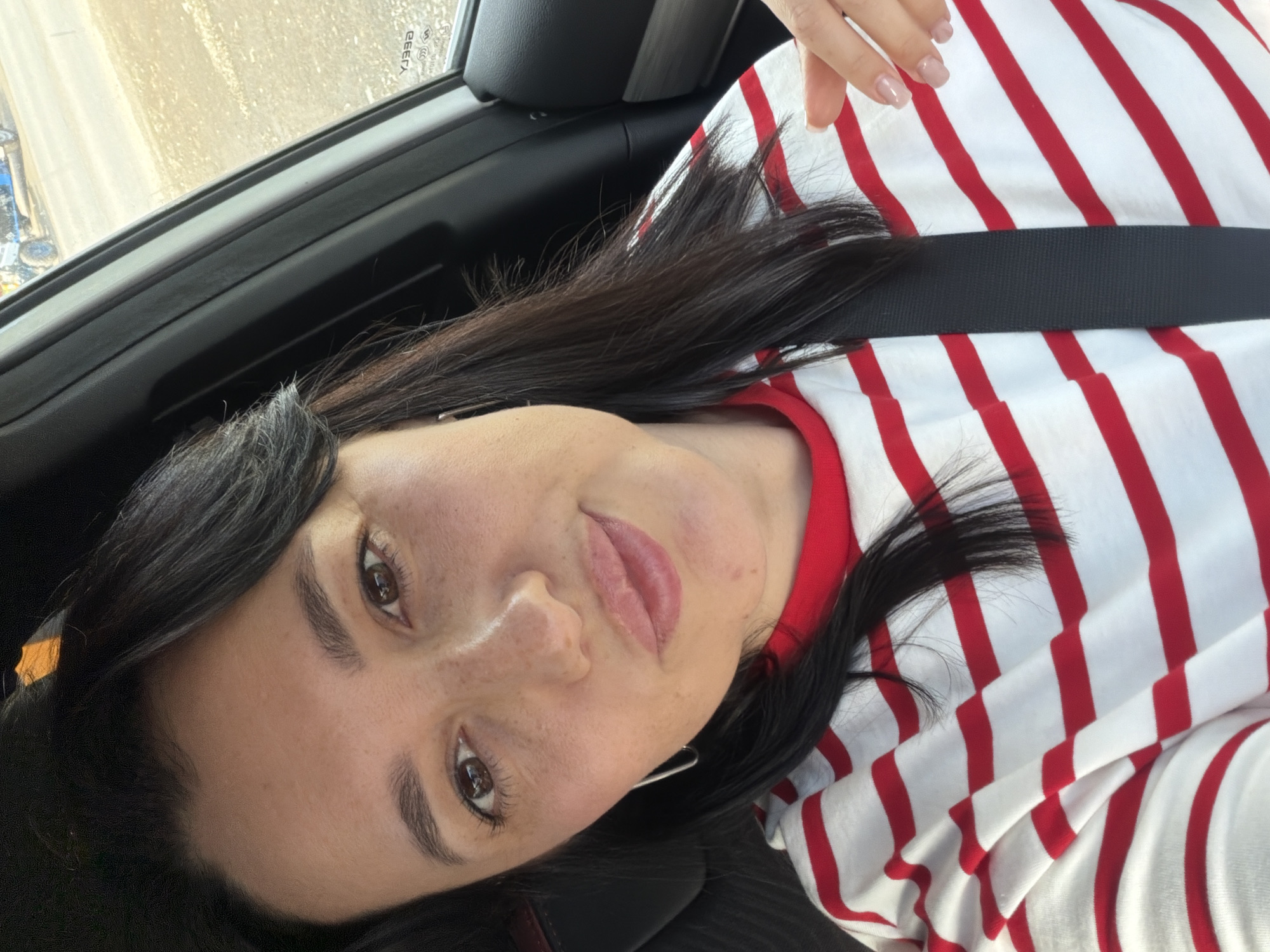 A woman with dark hair and fair skin is taking a selfie in a car. She is wearing a red and white striped shirt. Her hair is dark and long, falling around her shoulders. She has brown eyes and fair skin with freckles. Her lips are full and appear to be lightly glossed. The background shows the interior of a car, with a seatbelt visible. Outside the window, a dusty road and some vehicles can be seen. The lighting suggests it is daytime. There is no visible indication of the specific location in the city of Шмидтово, Russia.