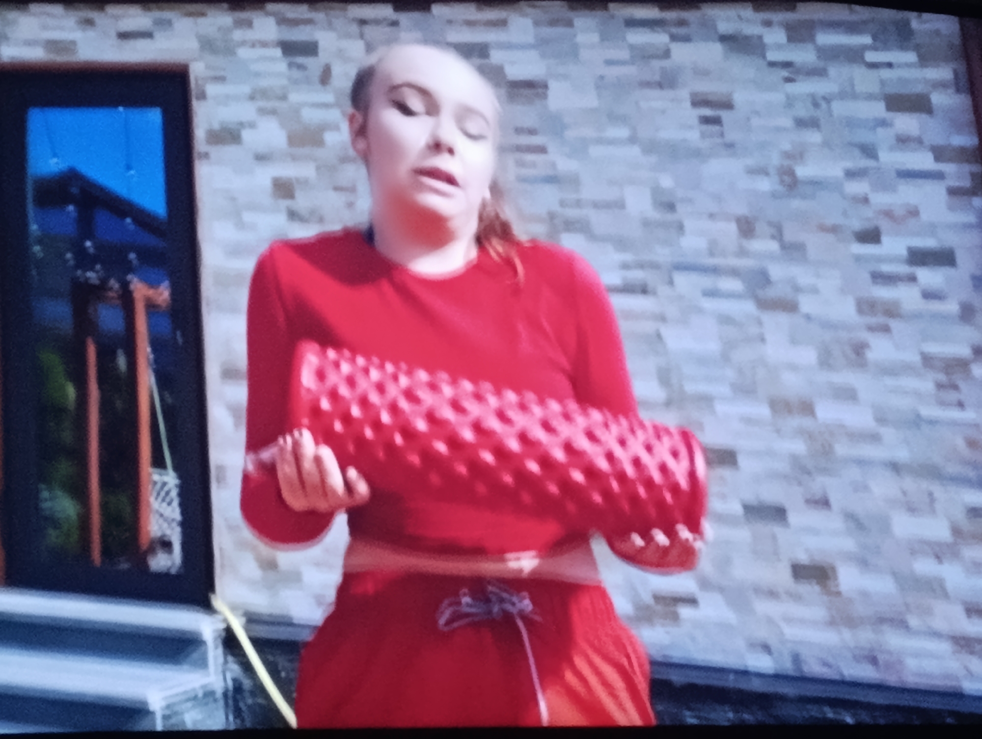 In a medium shot, a young woman stands outside a brick building, holding a red foam roller with a textured surface. She is wearing a red long-sleeved shirt and red shorts with a drawstring. Her eyes are closed, and her mouth is slightly open, suggesting she might be speaking or exhaling. The background features a wall constructed of light-colored rectangular bricks. To the left of the woman, a dark-colored doorway with glass panels is visible. The scene is likely a residential area in Dnipro, Ukraine, given the context.