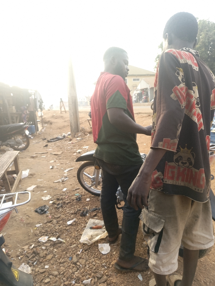Two people stand beside a motorbike in a dusty, litter-strewn environment. One wears a red and green shirt with dark pants and sandals, while the other sports a panda-printed shirt and light-colored shorts. The background shows a street with buildings and other people, suggesting a busy, possibly rural, area. The lighting is bright, washing out some details of the scene.